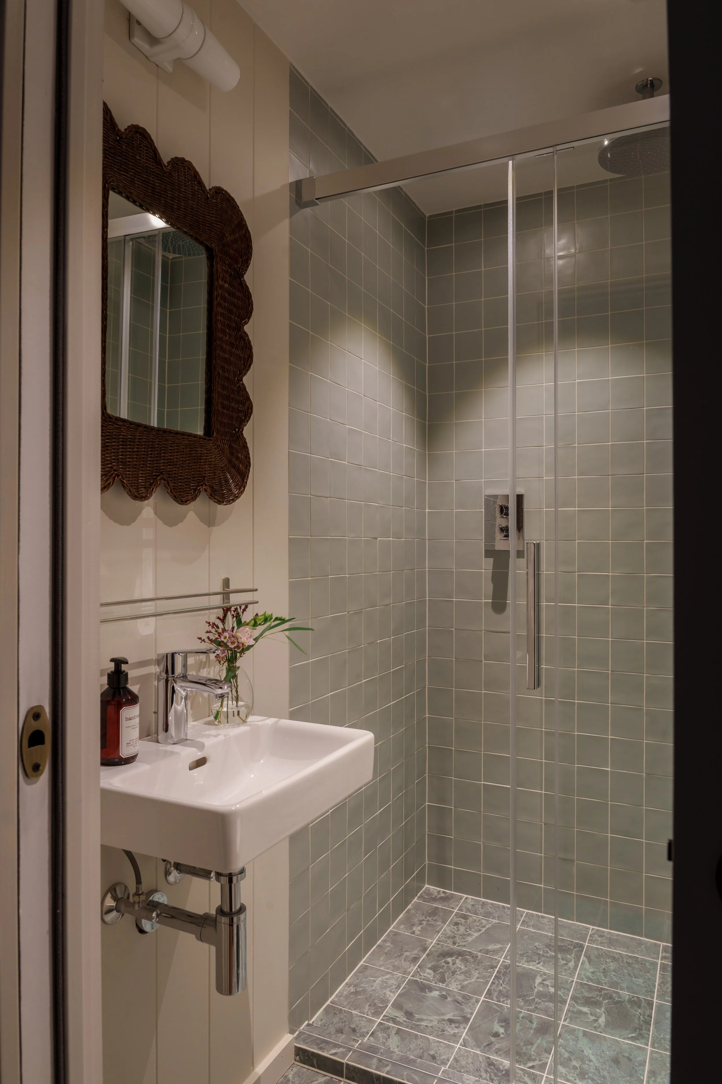 A small bathroom with a walk-in shower with glass sliding doors, a wall-mounted sink with a soap dispenser and a small plant, a mirror with a woven frame, and beige tiled walls and gray tiled floor.