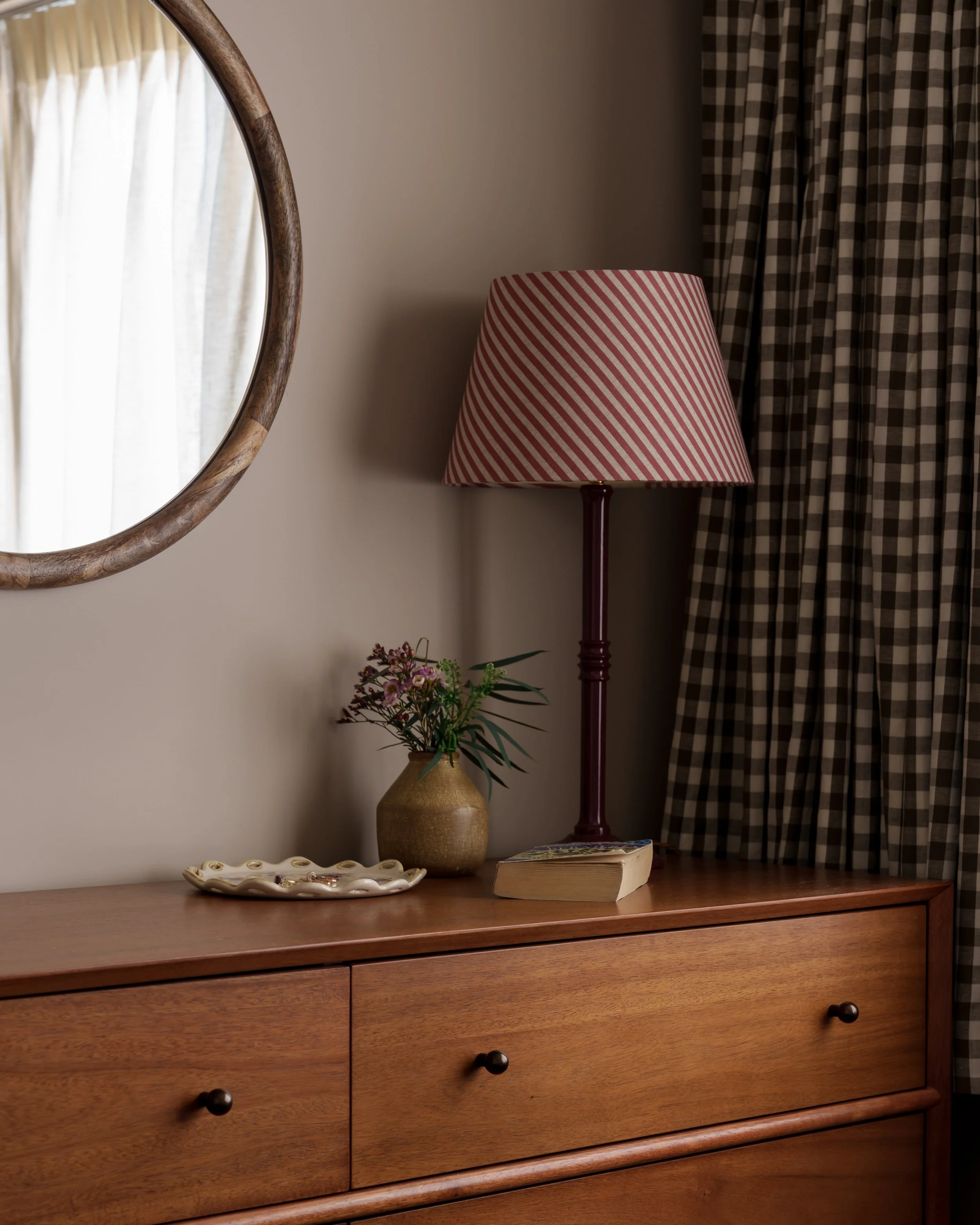 Wooden dresser with a vase, a book, a lamp with a pink-striped shade, and a decorative dish on top. A round mirror and checkered curtains are also visible.