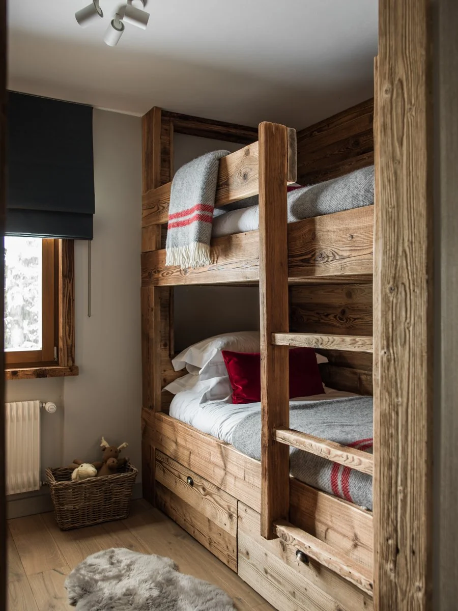 A cozy bedroom corner featuring a wooden bunk bed with two beds, one on top and one at the bottom, made of rustic wood. The top bed has a gray blanket with red stripes, and the bottom bed has white bedding with red pillows. A window with a dark blue 