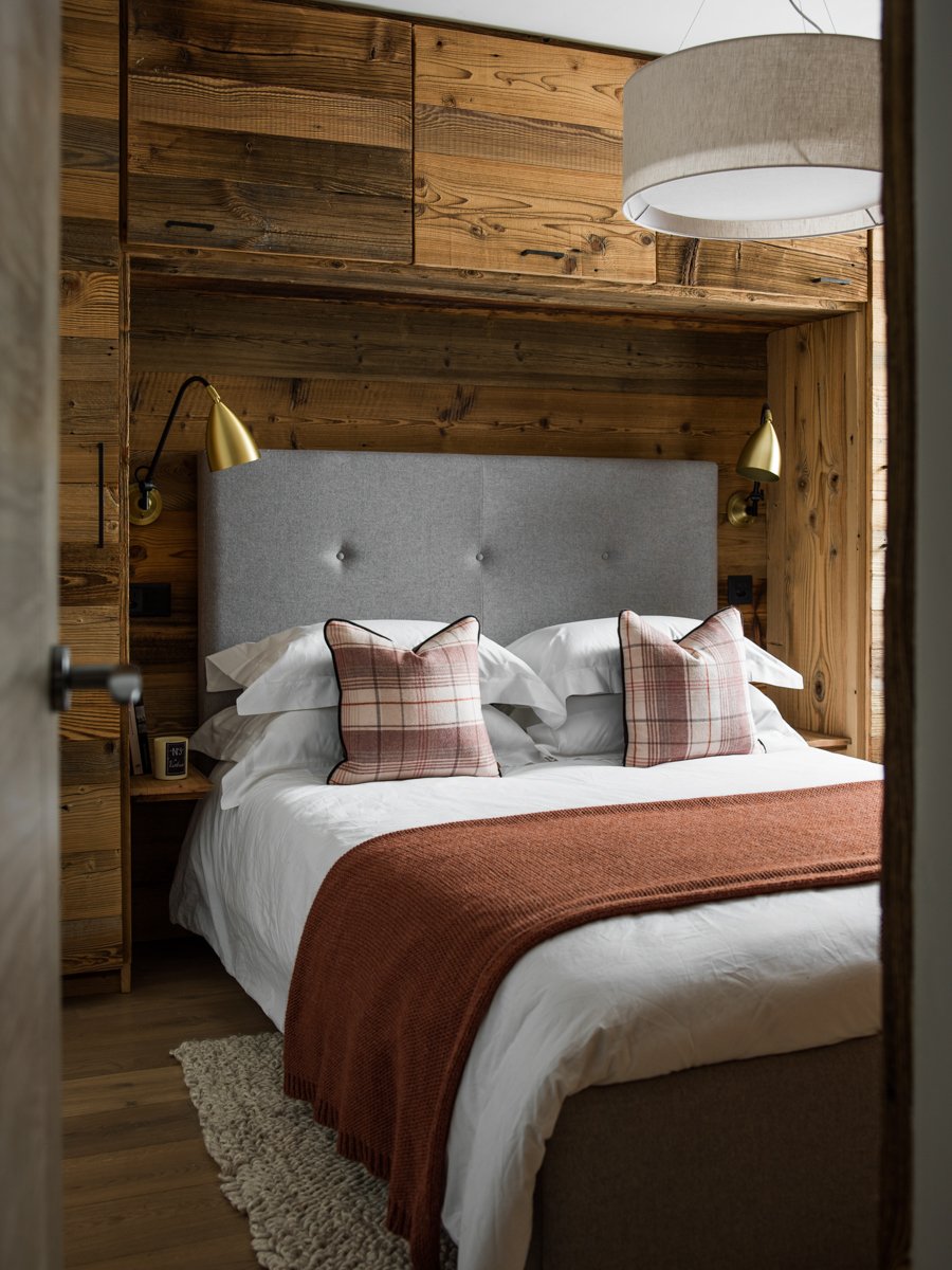 A cozy bedroom with a bed featuring a gray upholstered headboard, white linens, two plaid pillows, and a rust-colored throw blanket. The room has wood-paneled walls, wall-mounted gold reading lamps, a soft beige rug, and a small bedside table.