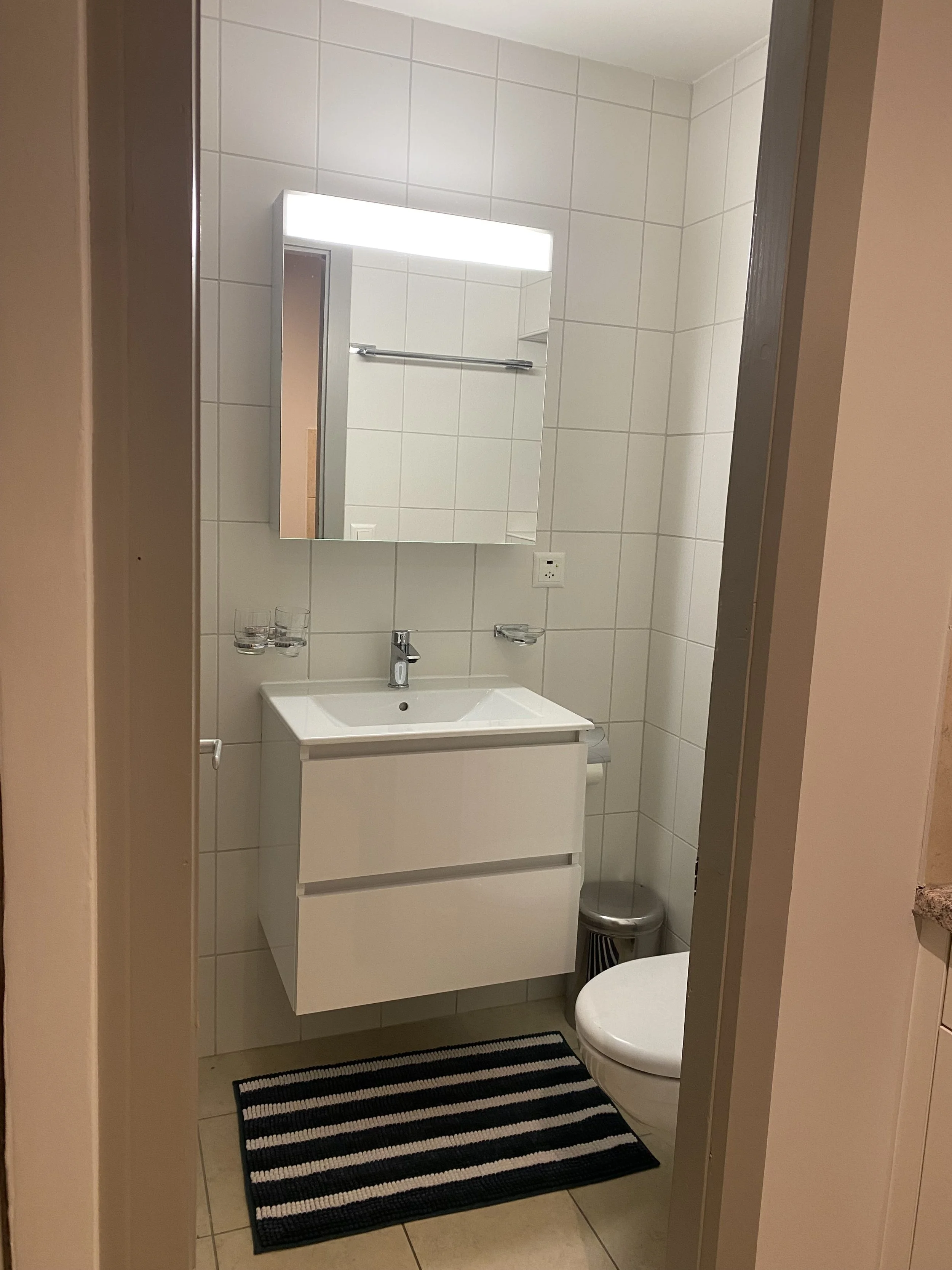 A small bathroom with white tiled walls and floor. It features a white vanity with a sink, a rectangular mirror above, a small trash can, a black and white striped rug, and a wall-mounted light strip above the mirror.