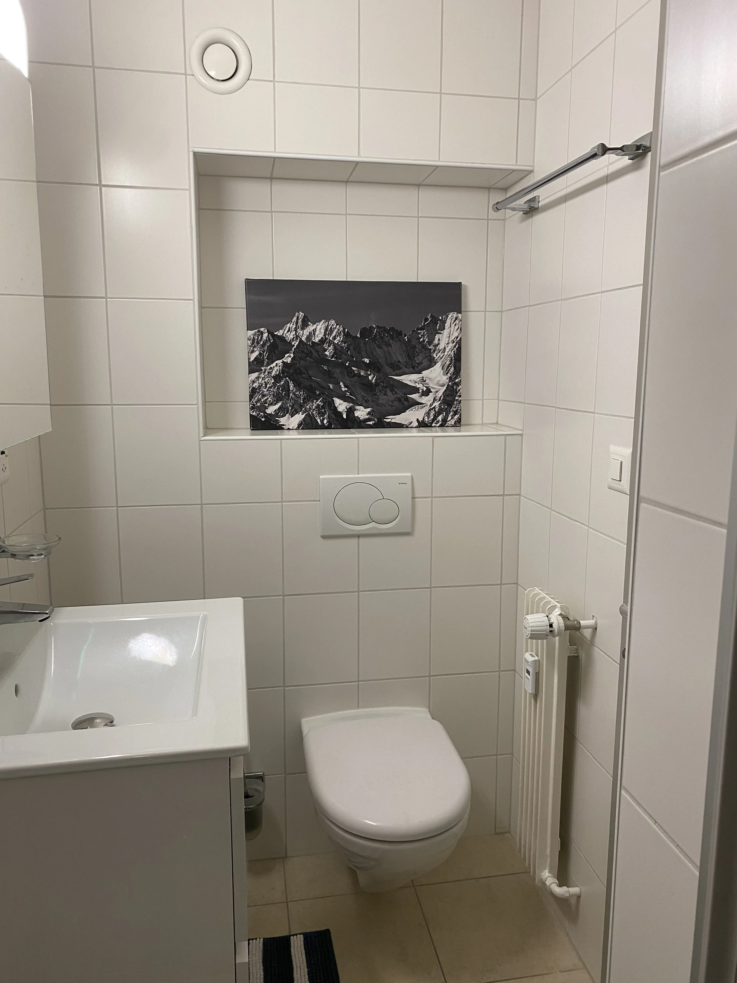 Small bathroom with white tiled walls and beige floor. Contains a white toilet, a small sink with a cabinet, a radiator with a thermostat, a black and white mountain artwork on a niche in the wall, and a towel rail. There is a vent in the upper wall.