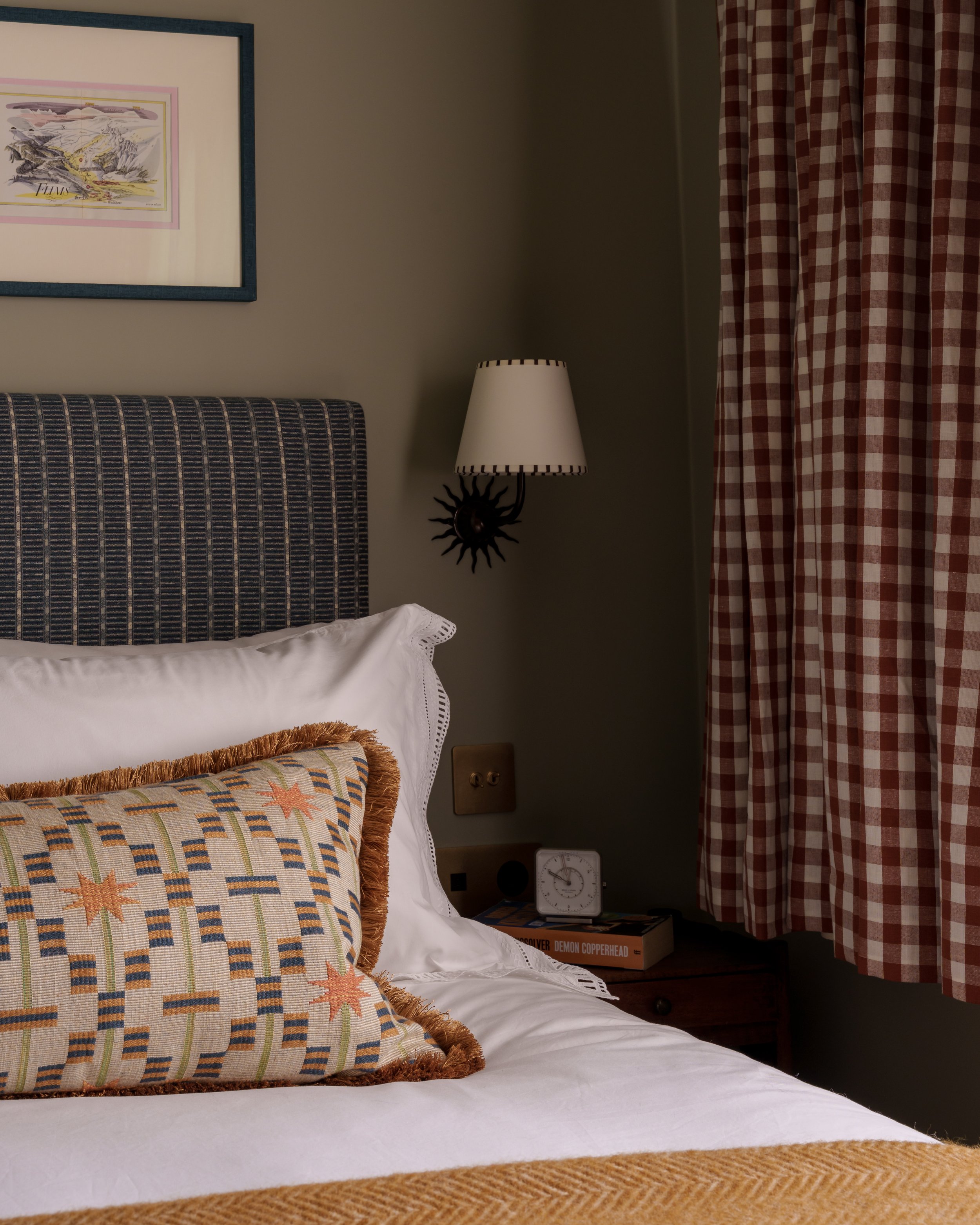 A cozy bedroom corner with a bed, embroidered pillow, wall art, bedside lamp, clock, and books near checkered curtains.