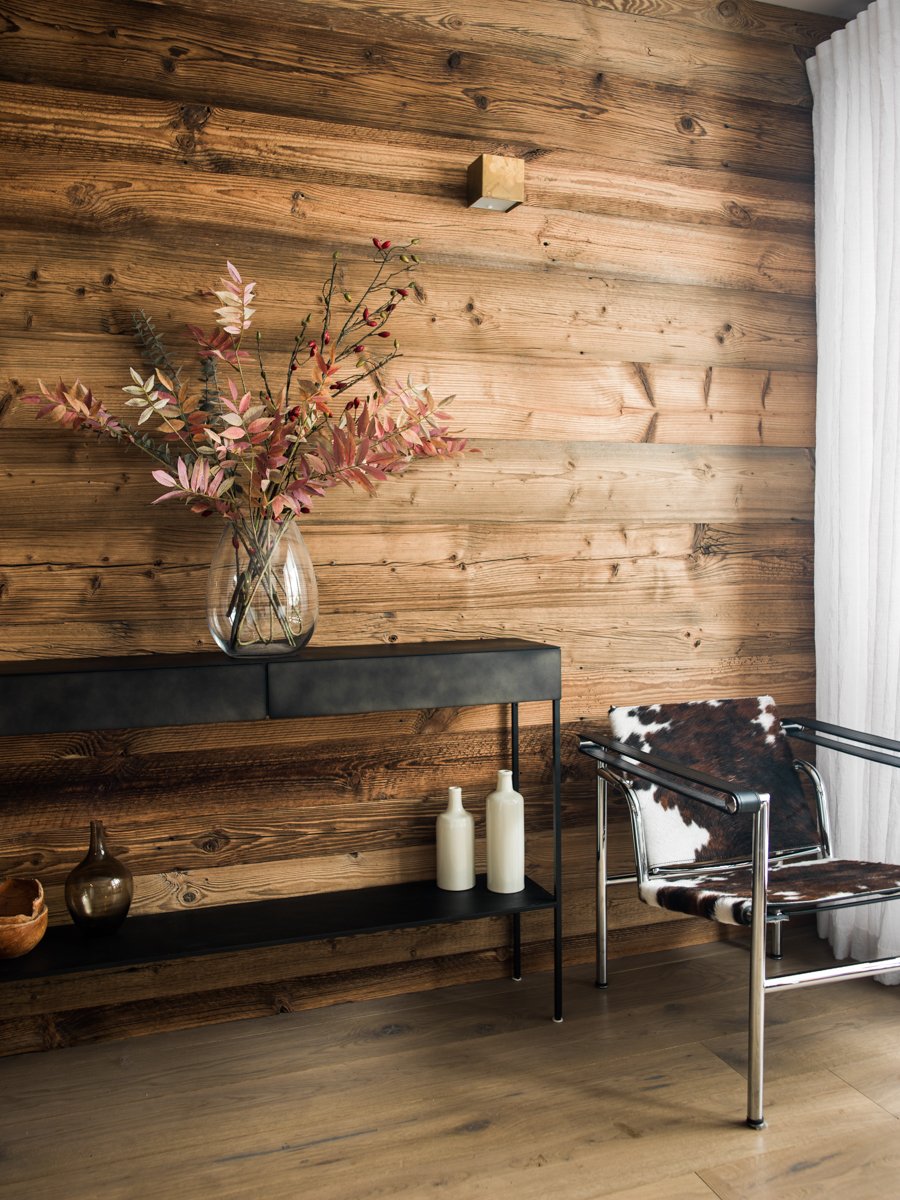 Interior view of a room with wood-paneled wall, a black metal console table with decorative vases, a chair with cowhide upholstery, and sheer white curtains.