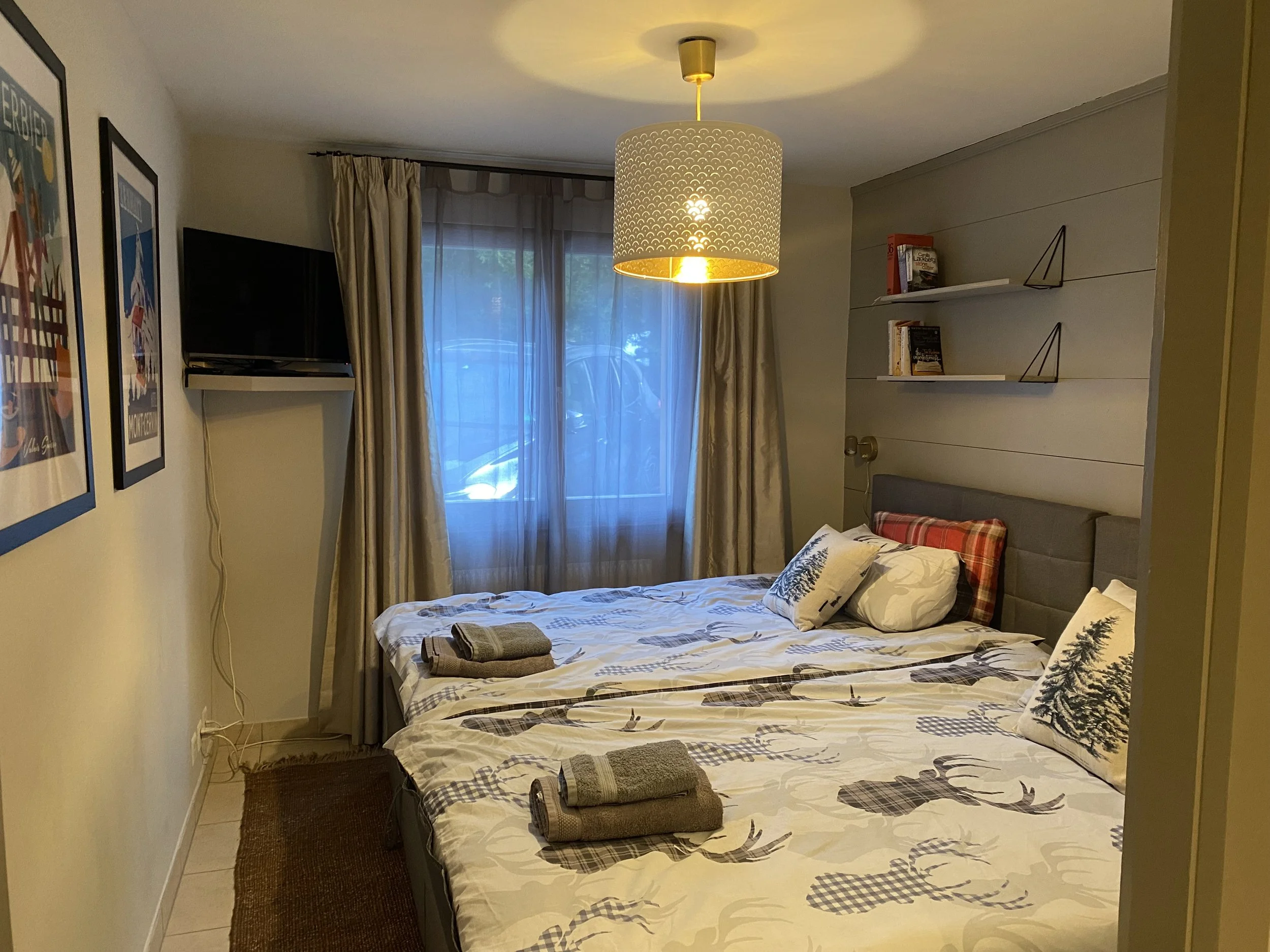 A cozy bedroom with two beds decorated with reindeer and tree-themed bedding, pillows, and blankets. There is a small window with curtains, a mounted TV on the wall, and a ceiling light fixture. Shelves with books are on the wall above the beds.