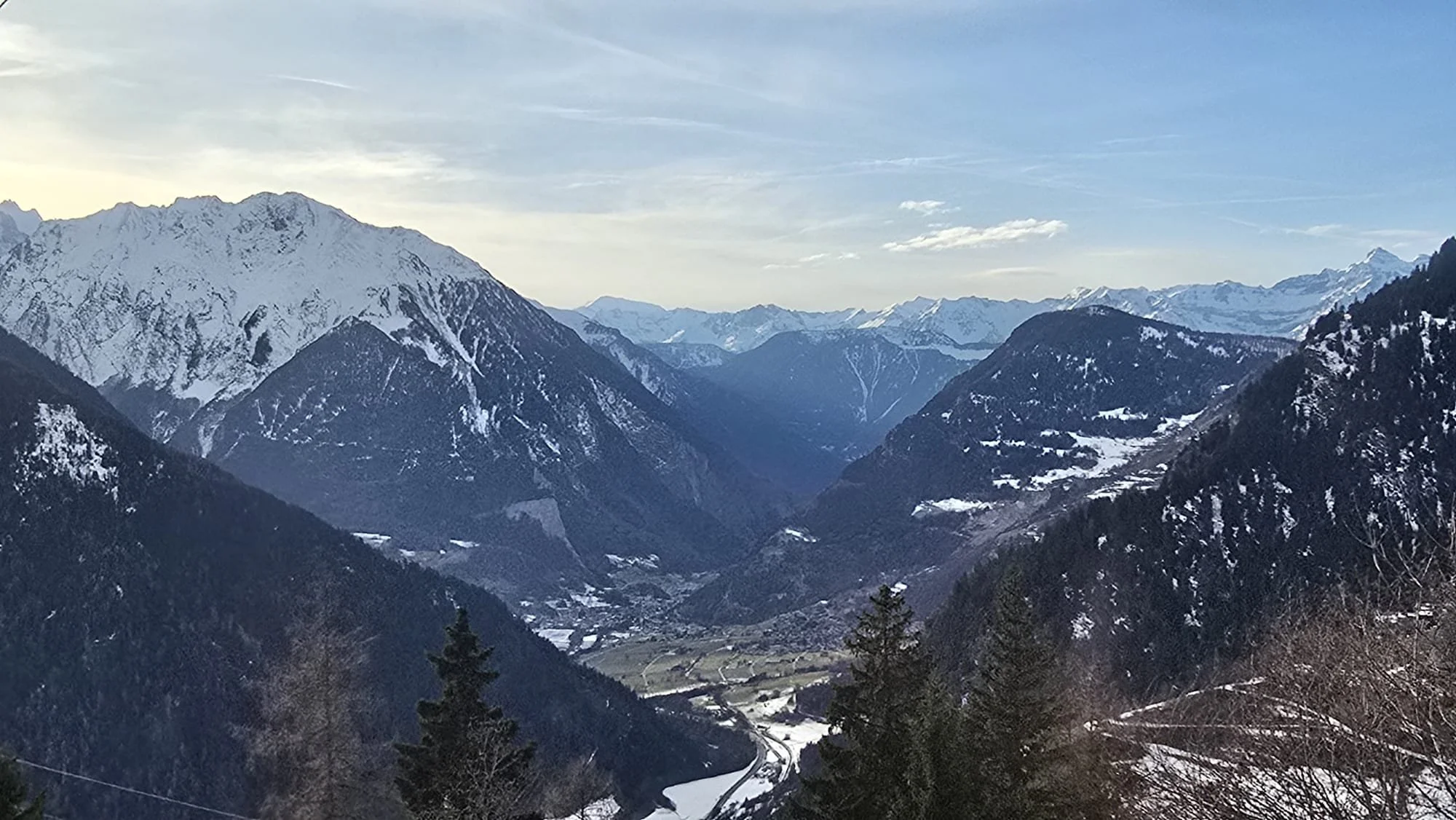 Snow-capped mountains in a valley with a winding river, trees in the foreground, under a partly cloudy sky.