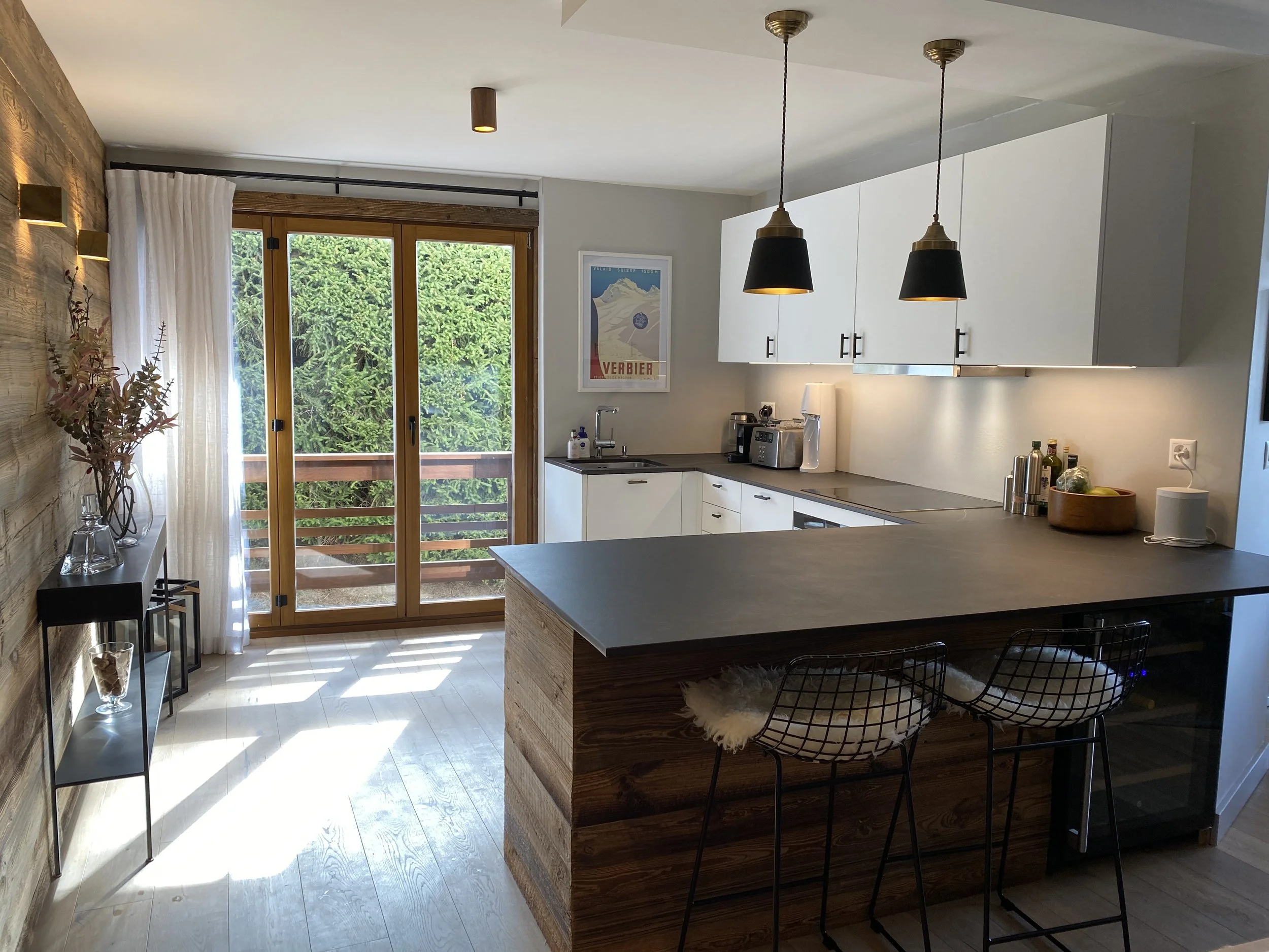 Modern kitchen with wooden accents, white cabinets, and a sliding glass door leading to a balcony with greenery. Includes barstools, a countertop with various appliances and decor, and a wood-paneled wall with decorative items.