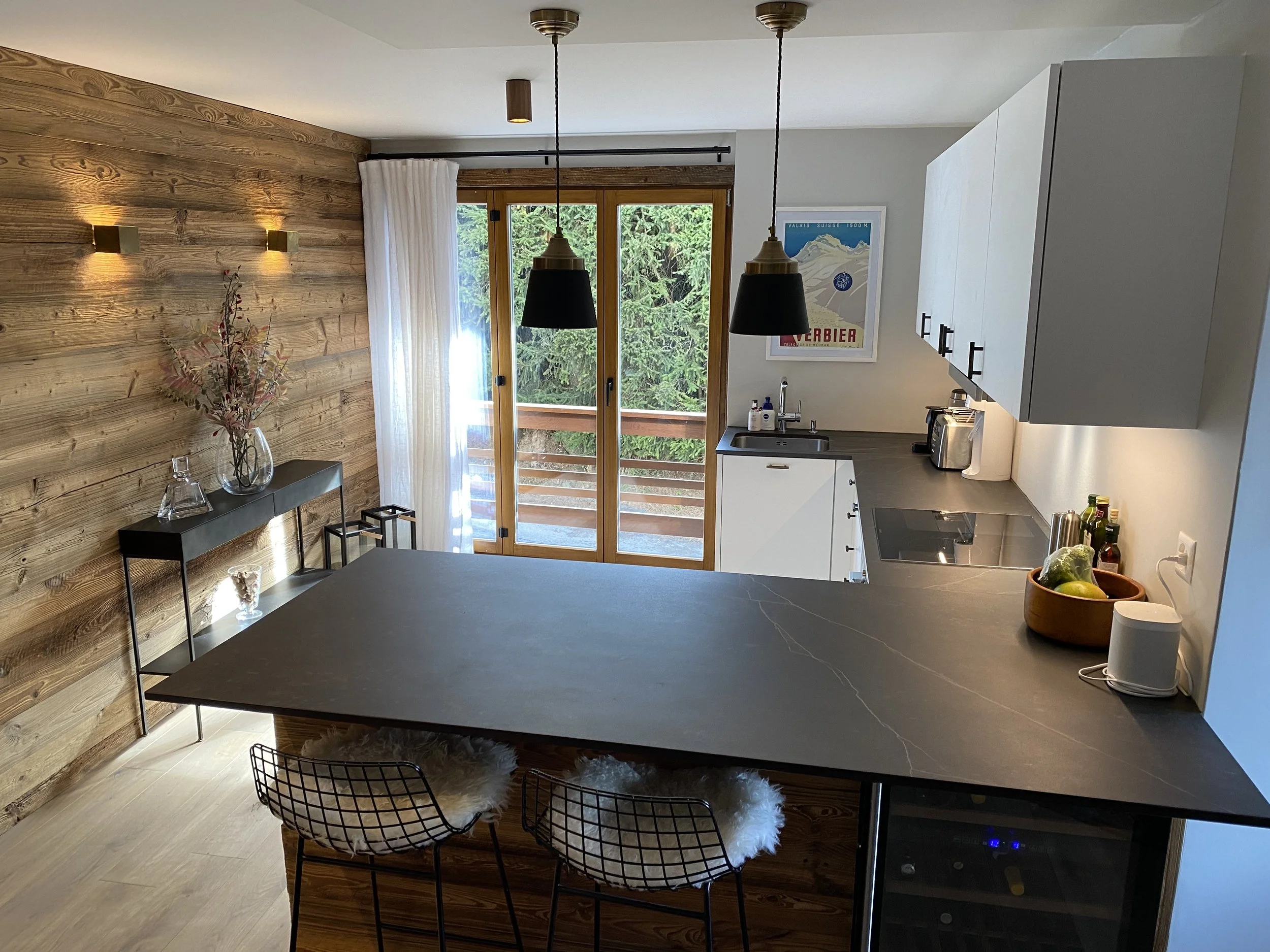 Modern kitchen with wooden accent wall, black pendant lights, white cabinetry, and a sliding glass door leading to a balcony, overlooking greenery.