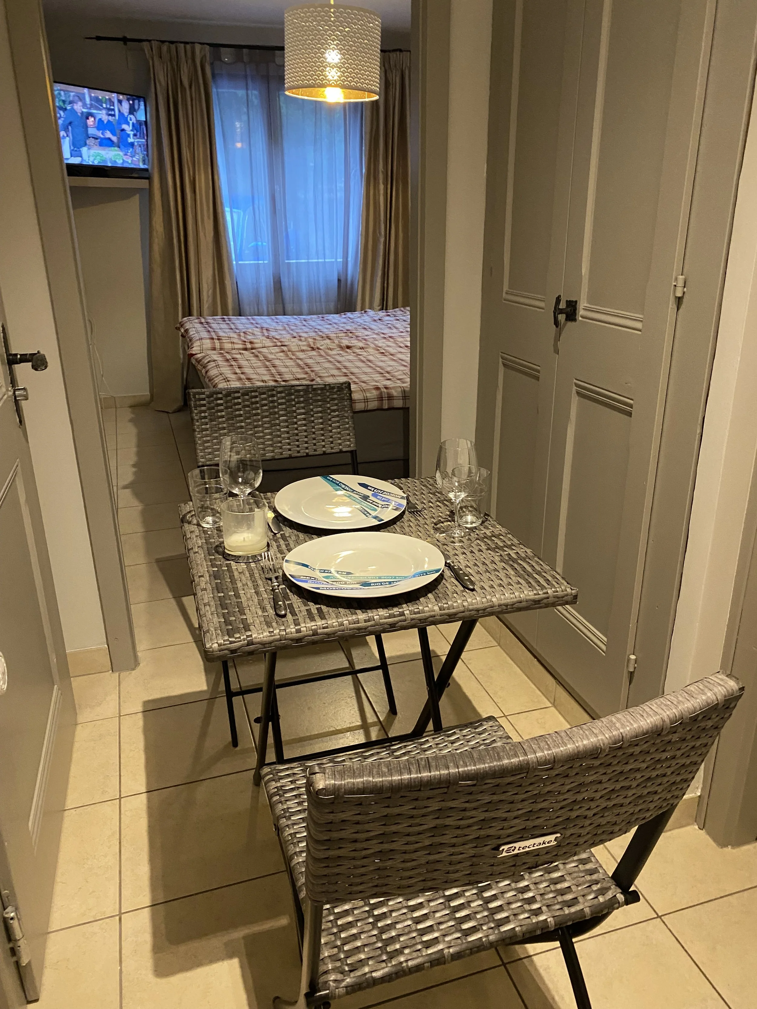A small dining area with a woven table set for two, including plates, glasses, and utensils. In the background, a bedroom with a bed covered in a checked bedspread and a window with curtains. A small TV is mounted in the corner.
