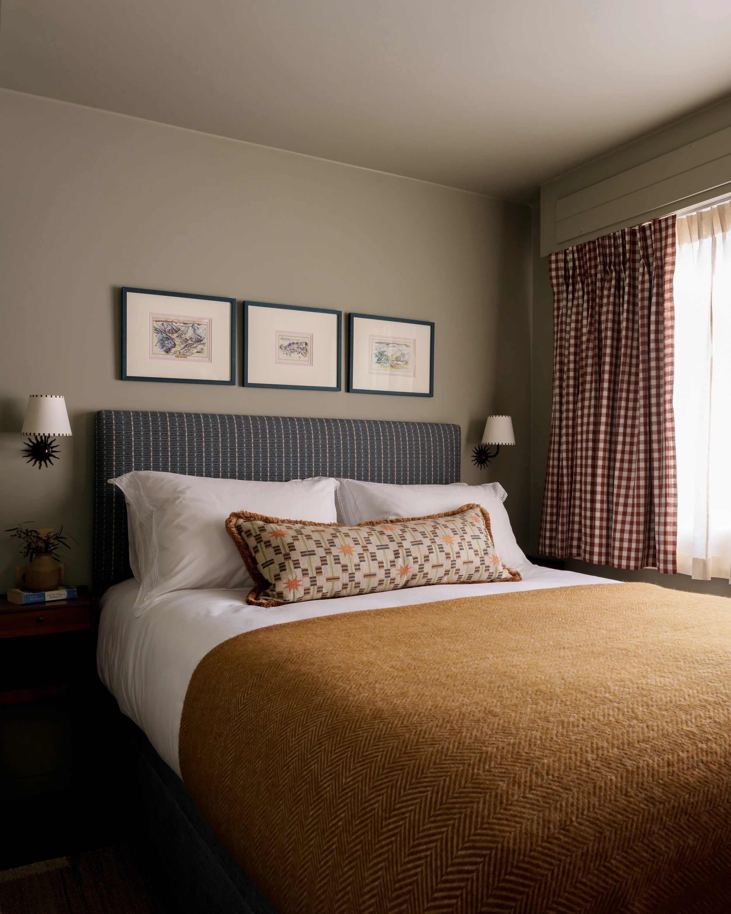 A neatly made bed with white pillows, a decorative pillow, and an orange-brown blanket in a cozy bedroom. There are three framed pictures above the bed and matching wall sconces on either side.