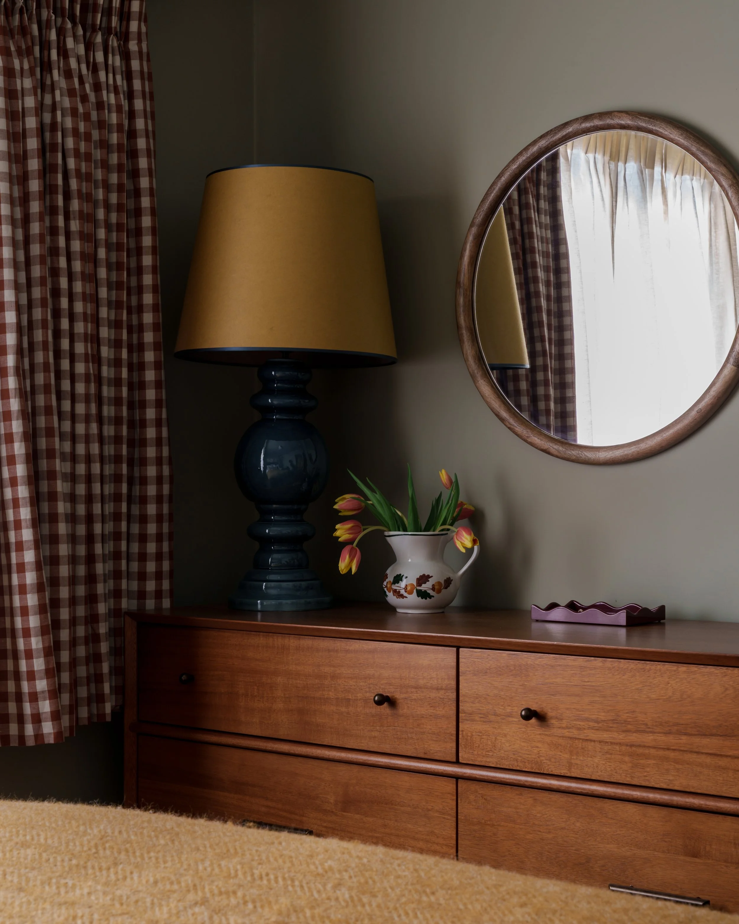 A wooden dresser with four drawers, a large black lamp with a yellow shade, a vase with yellow tulips, and a small decorative dish on top. A round mirror is mounted on the wall, and a window with checkered curtains is reflected in the mirror.