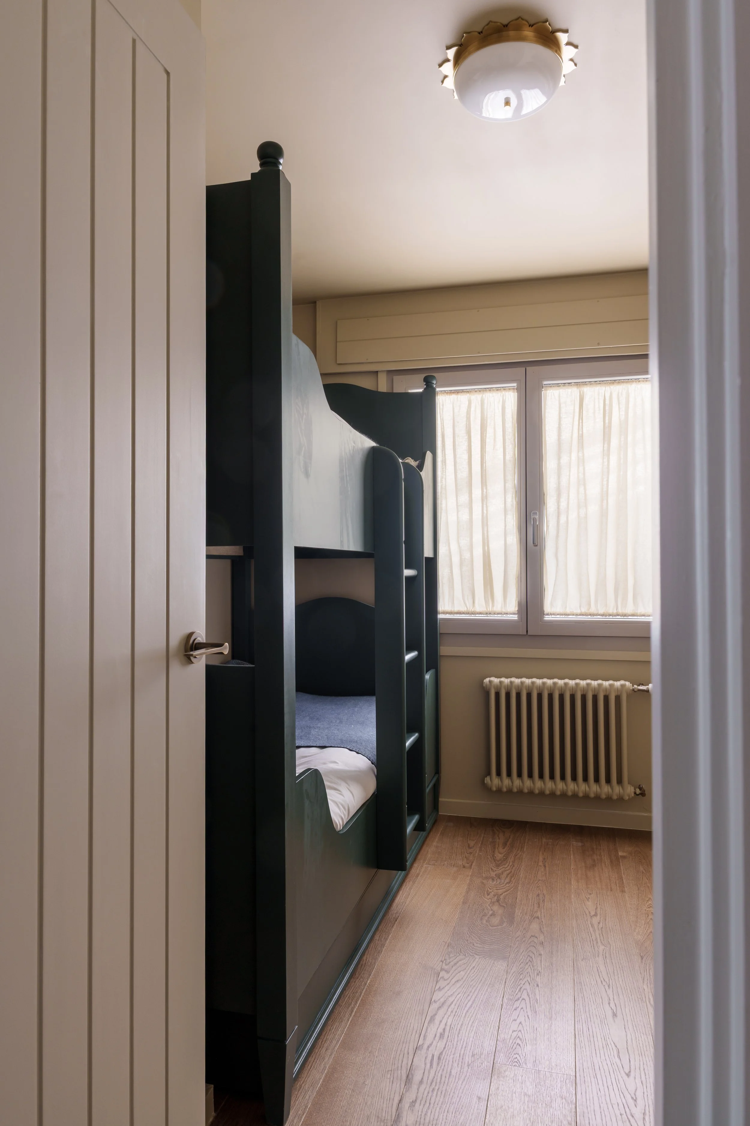 View of a small bedroom with a black bunk bed placed near the window, beige curtains, a white radiator, and a wooden floor. The photo is taken from the doorway.