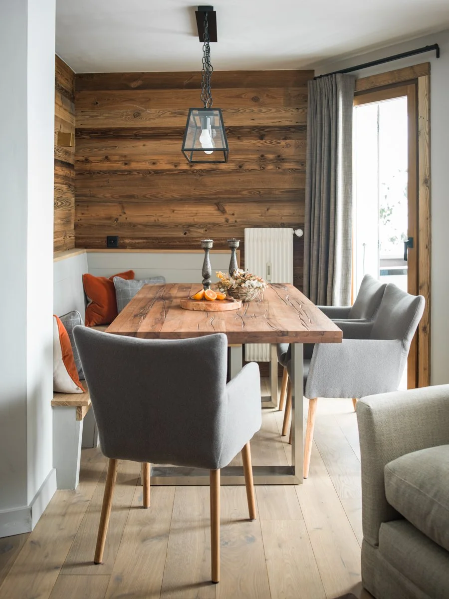 A dining room with a wooden table, gray chairs, and a wall of wooden planks. There are candles, a basket with oranges, and cushions on a built-in bench. A large window with curtains and a hanging light fixture are also visible.