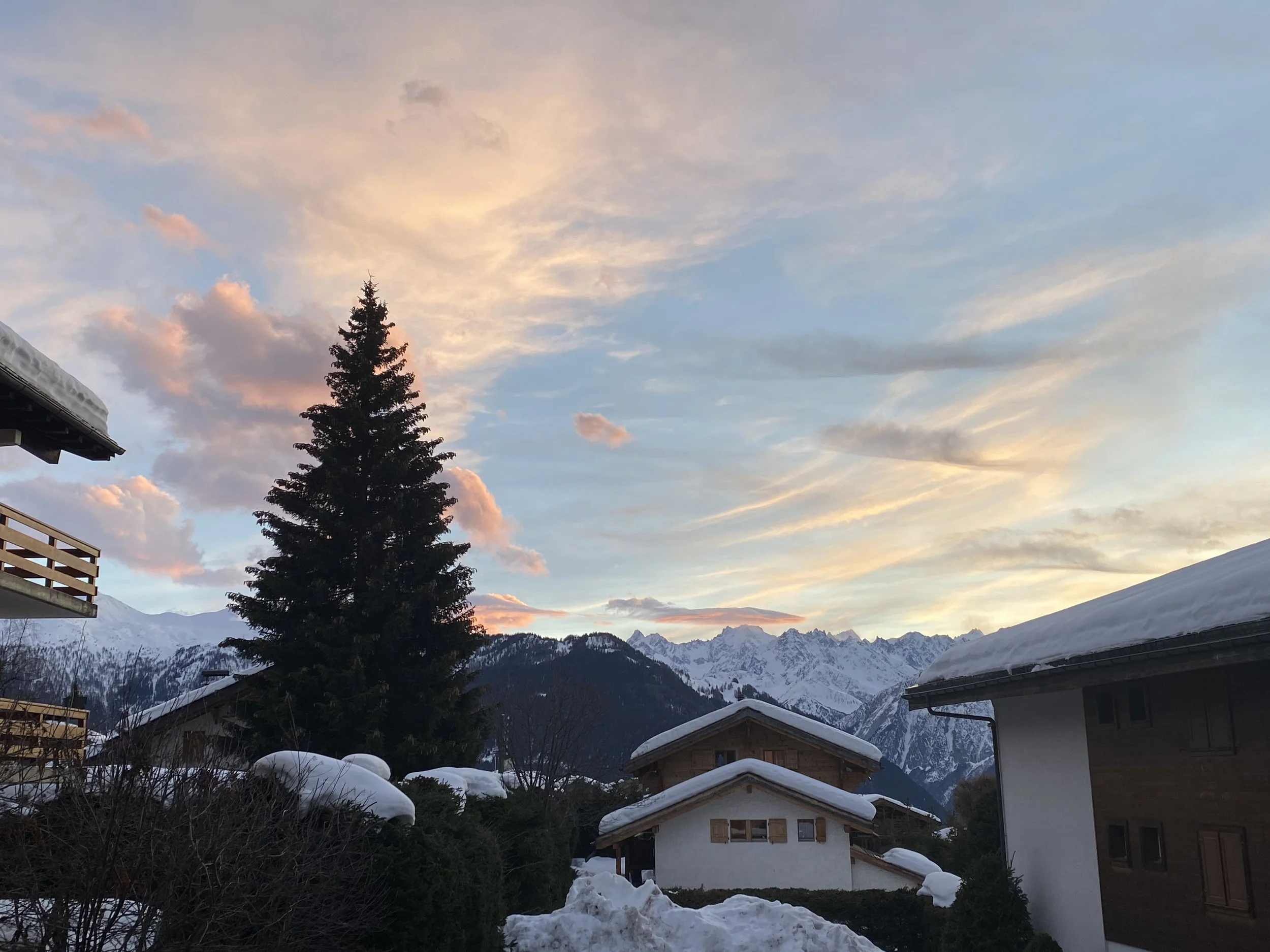 Snow-covered houses and trees against a backdrop of mountains under a colorful sky at sunset.