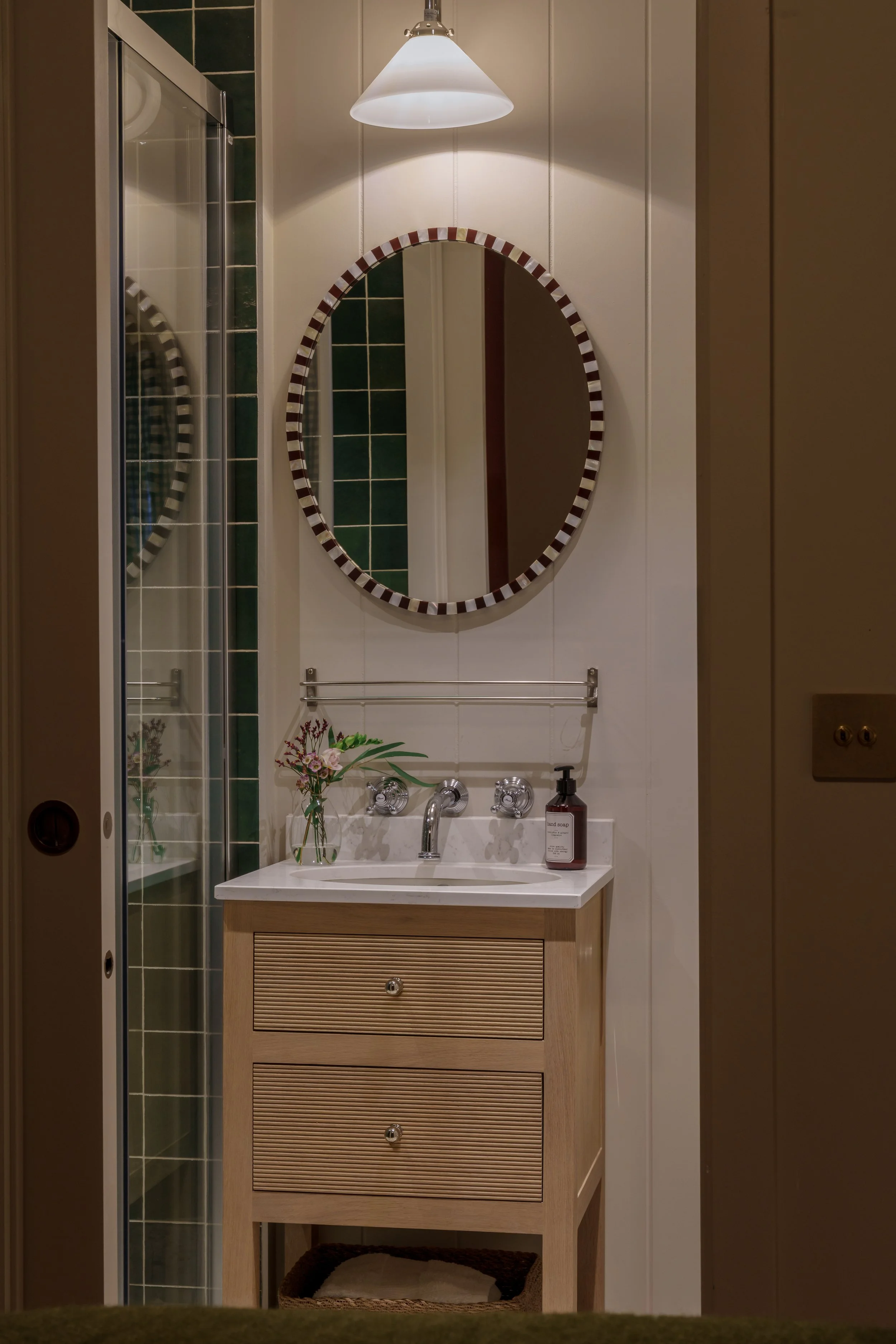 A small bathroom with a white wall-mounted sink, a round mirror with a striped frame, and a flower arrangement on the sink. There is a glass shower door to the left and a light fixture above the mirror.