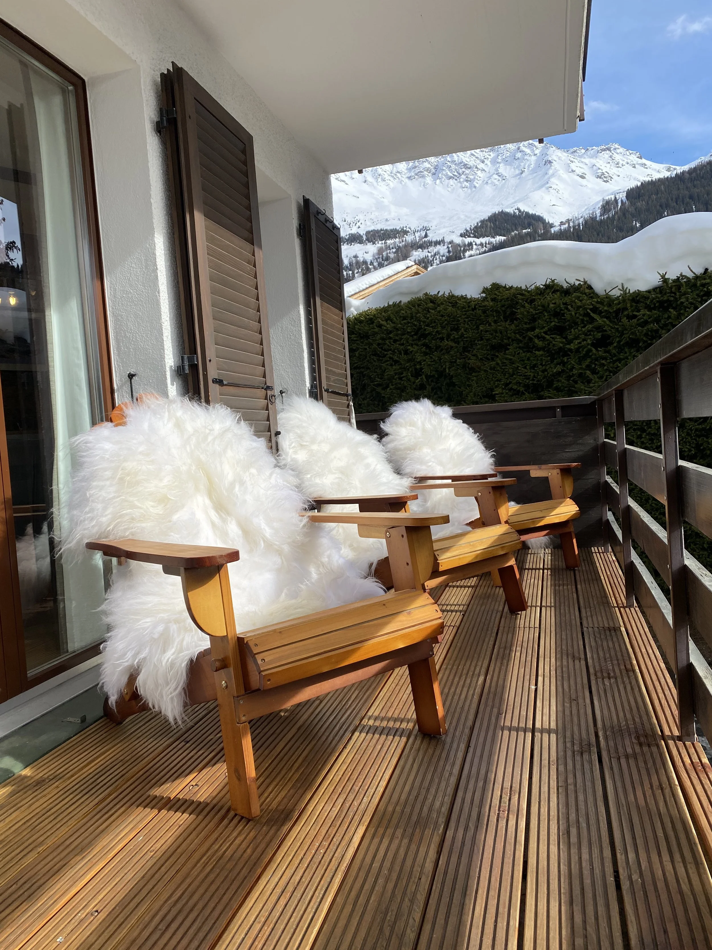 Three wooden chairs with white fur throws on a balcony overlooking snow-covered mountains and green trees.