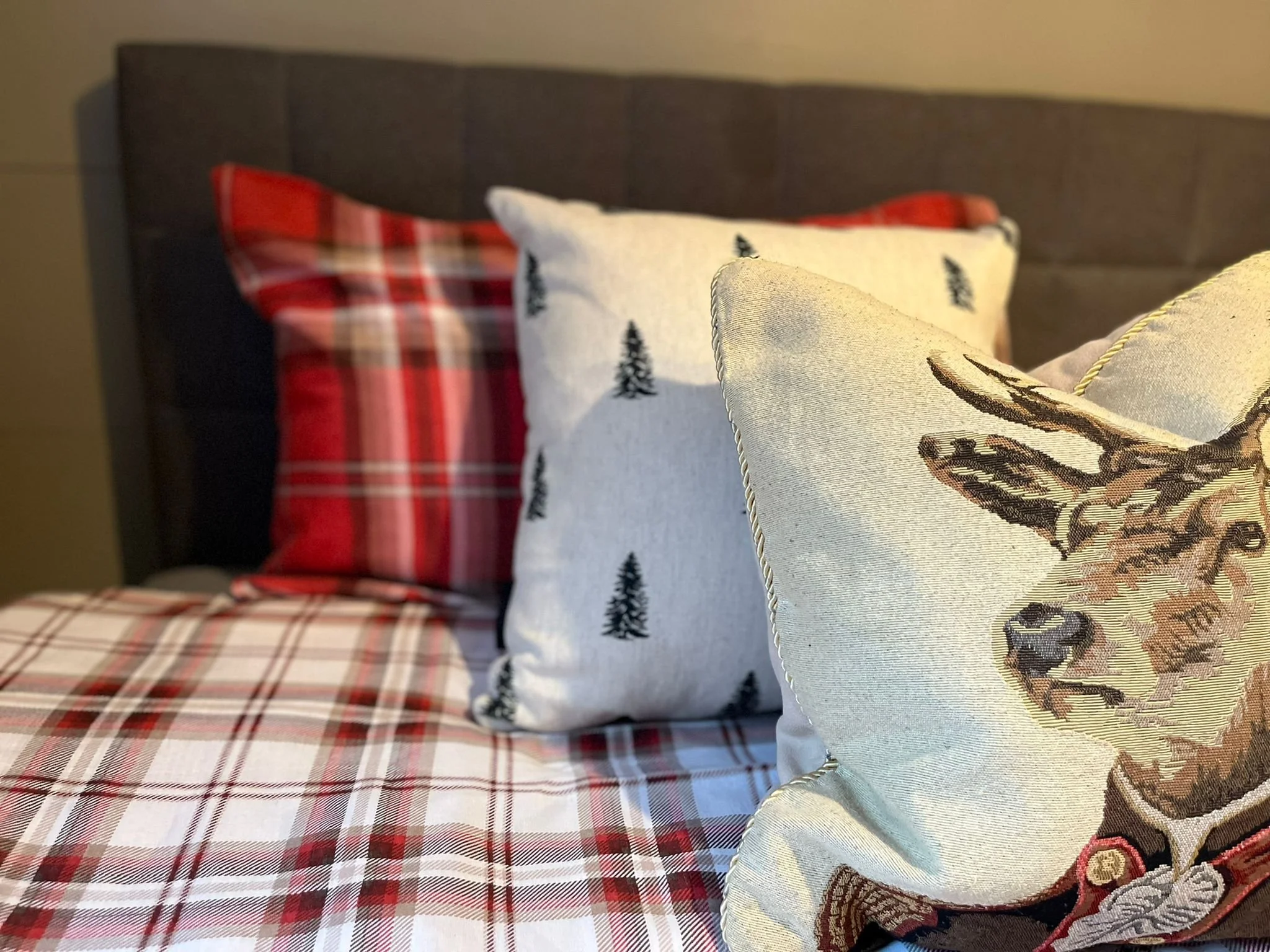 A bed with red and white plaid bedding, a dark headboard, and three decorative pillows: one with a white background and small pine trees, another with a red plaid pattern, and a third with a deer head design.