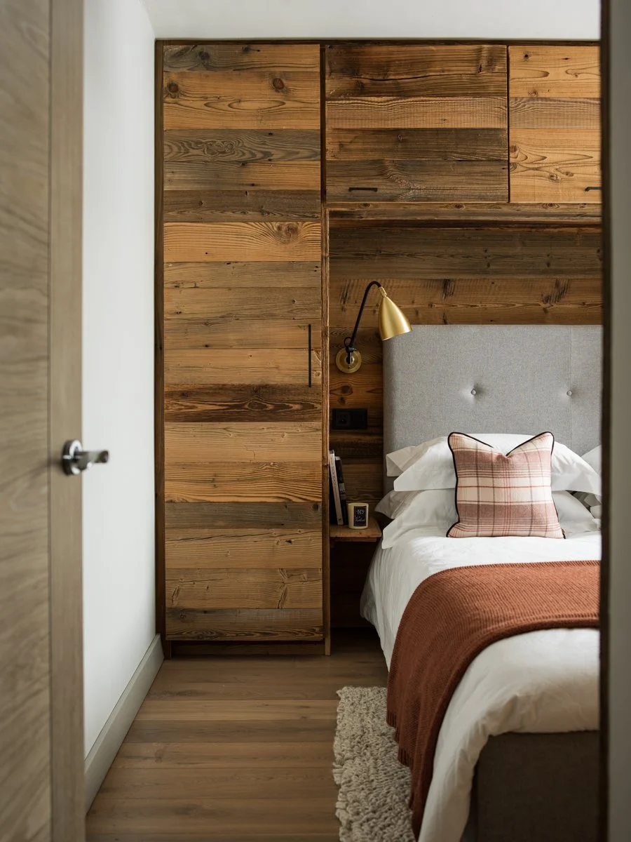 A cozy bedroom with a wooden paneled wall, a bed with white linens and a plaid pillow, and a gold reading lamp mounted on the wall.