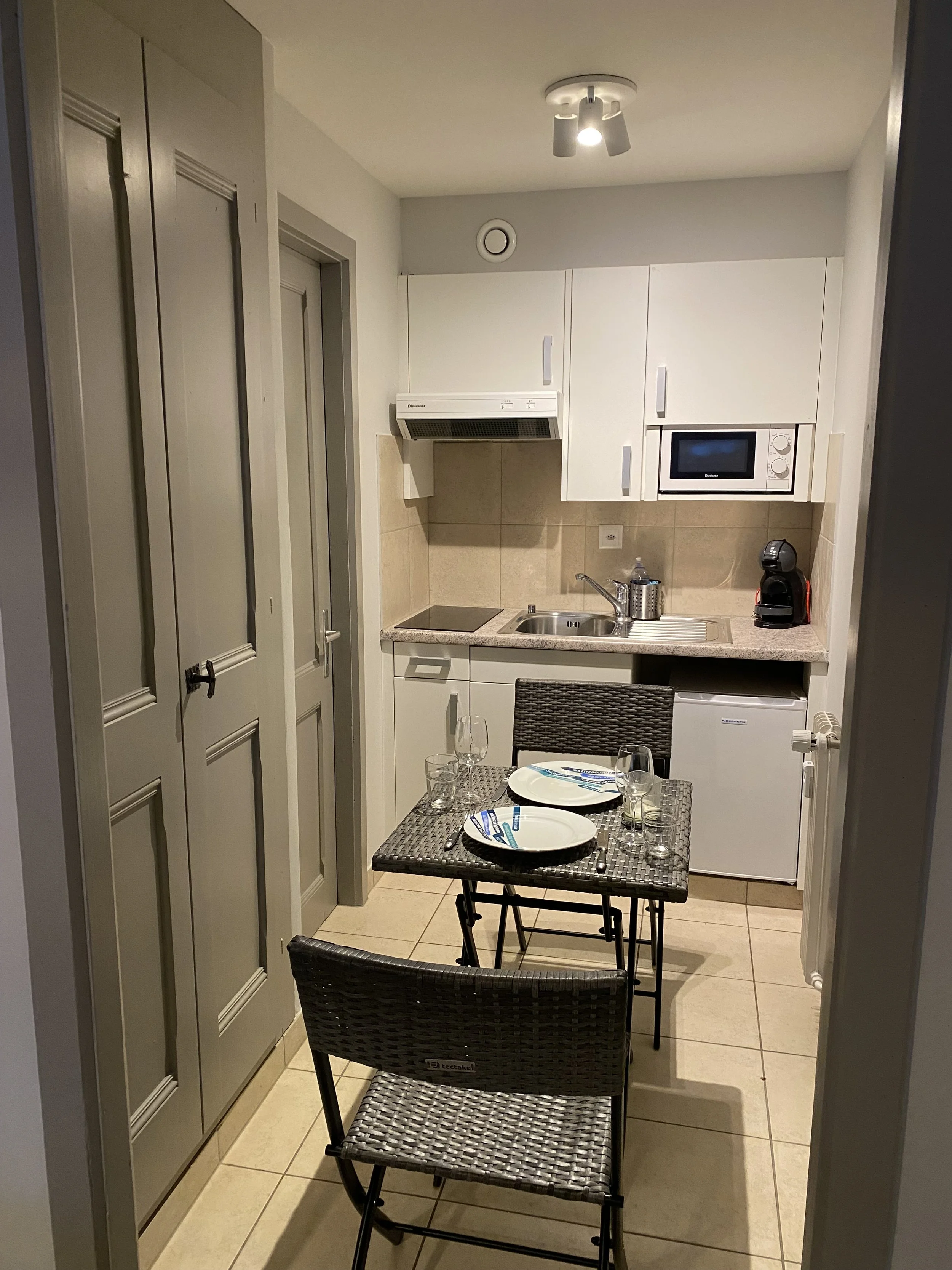 Small kitchen with white cabinets, microwave, coffee maker, small refrigerator, sink, stovetop, and a dining table set for two with plates, glasses, and utensils.