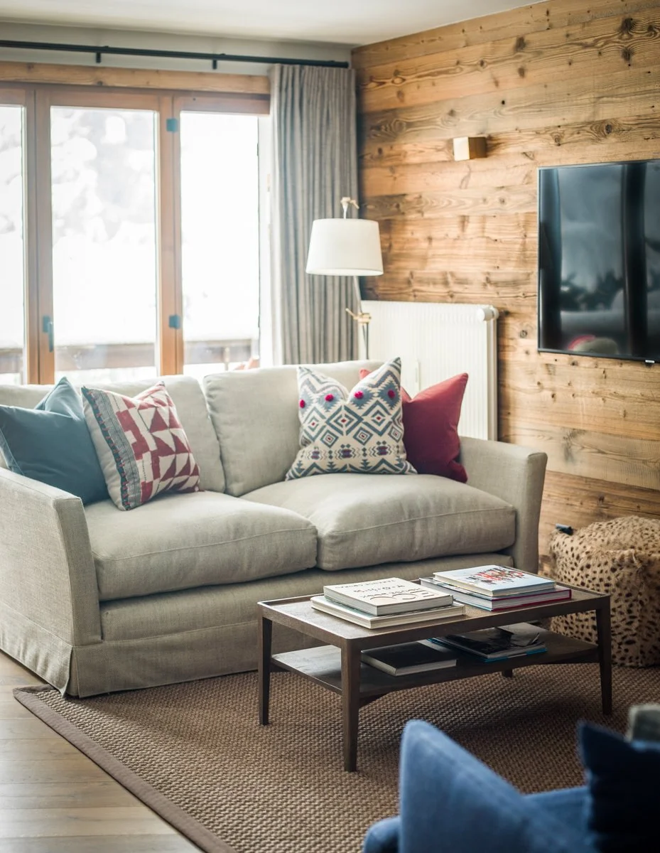 Living room with a gray sofa, decorative pillows, a wooden coffee table with magazines, a wooden accent wall with a mounted TV, a floor lamp, a radiator, and large windows with curtains.