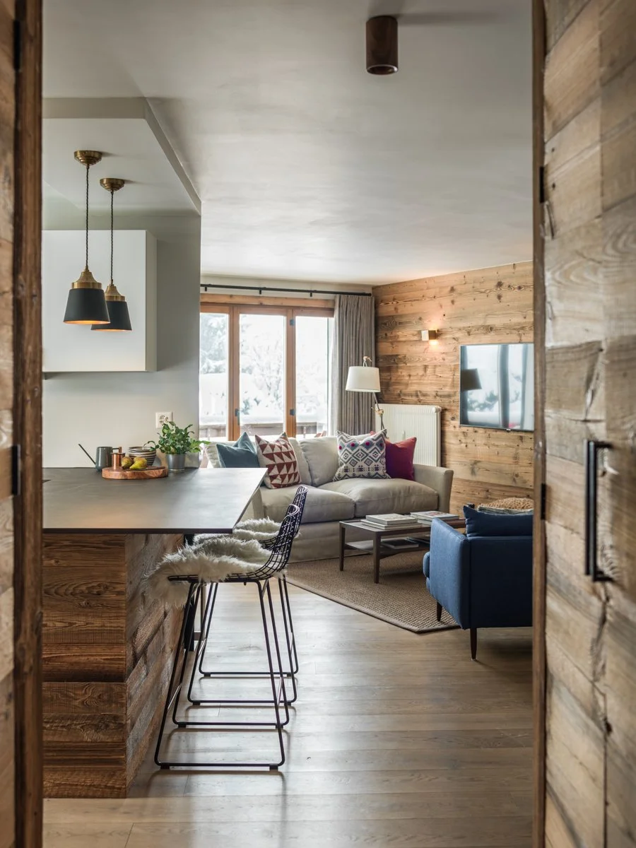 A cozy living room with wooden accents, a white sofa with patterned pillows, a blue armchair, a mounted flat-screen TV, a large window, and modern light fixtures.