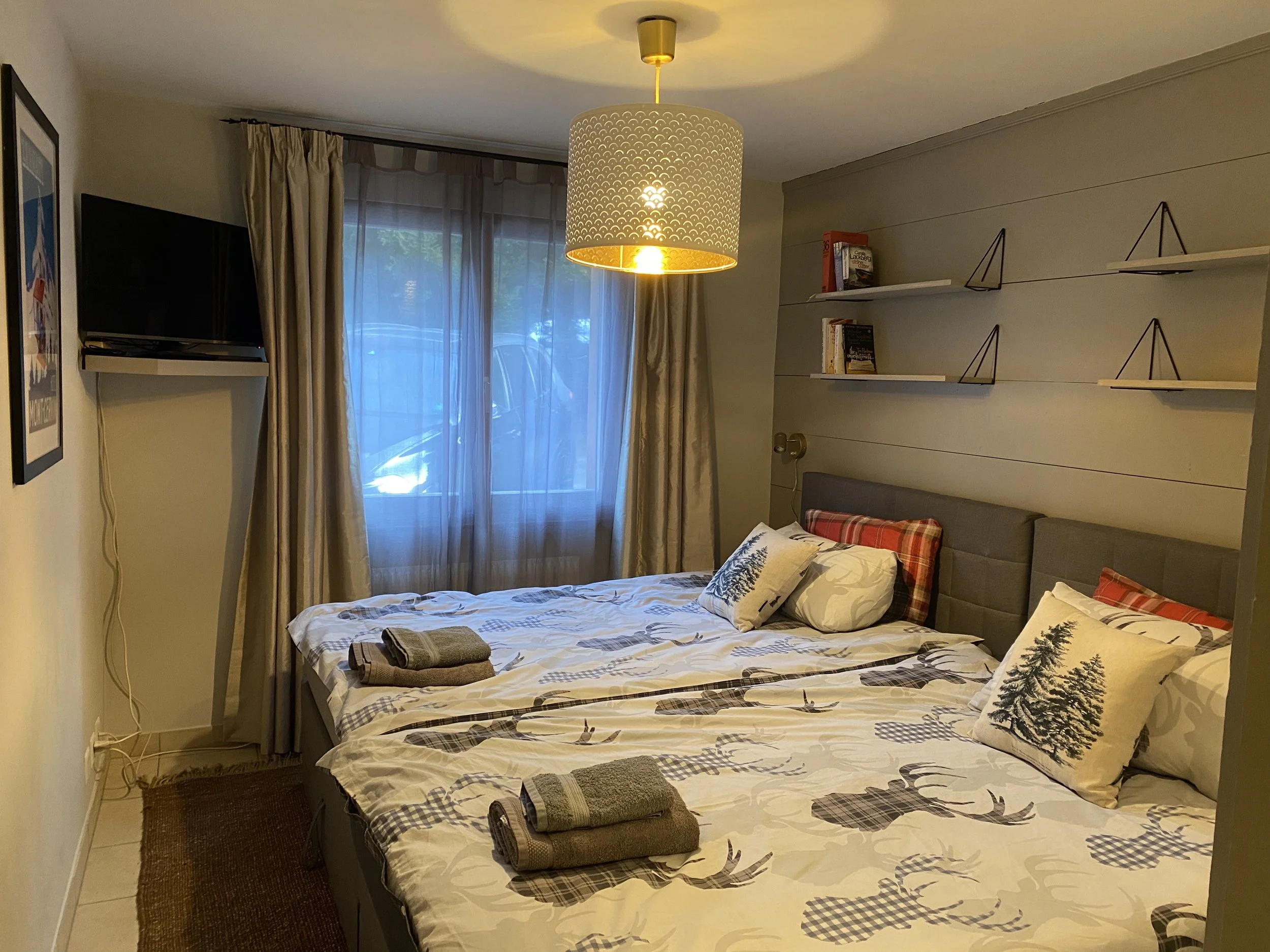 A bedroom with a bed, pillows with pine tree and plaid patterns, towels on the bed, a window with beige curtains, a wall-mounted TV, a ceiling light, and a wall shelf with books.