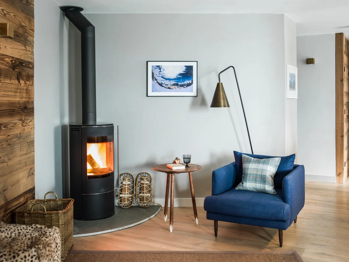 Living room with a wood-burning stove, a small wooden side table with a book and glass, a blue armchair with a plaid pillow, a gold floor lamp, framed artwork, and natural wood accents.