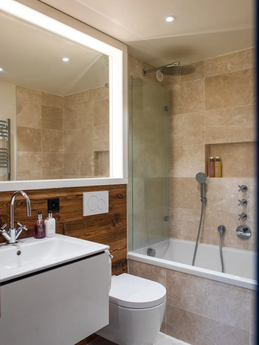 Modern bathroom with beige tiled walls, a white sink with a chrome faucet, a large mirror with integrated lighting, a wall-mounted toilet, and a bathtub with a glass shower screen, showerhead, and built-in niche for toiletries.