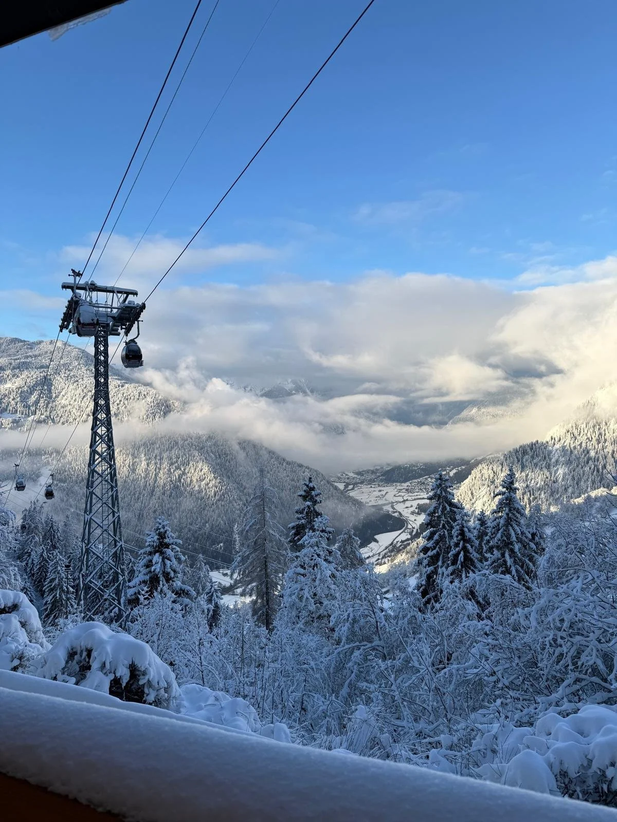 Snow-covered mountain landscape with pine trees, cable cars, and a valley below, under a partly cloudy blue sky.
