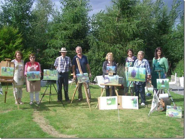 A group of eight people standing outdoors in front of trees, displaying their paintings of lakes and nature scenes on easels and canvases.