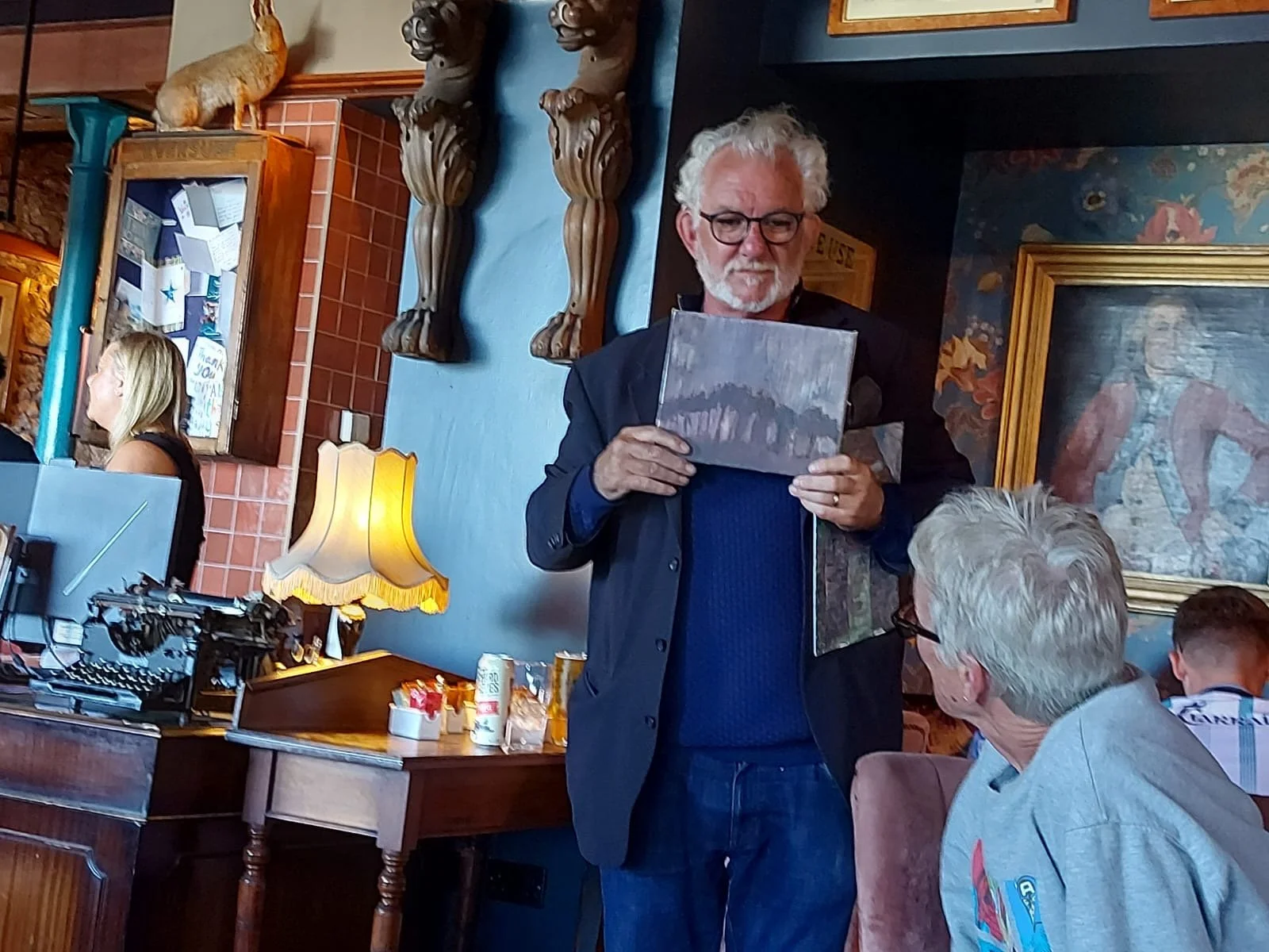 An older man with white hair and glasses stands inside a restaurant holding a painting, talking to a seated man with grey hair and glasses. The background features a blue wall with sculptures of lions, framed artwork, and a woman with blonde hair wor
