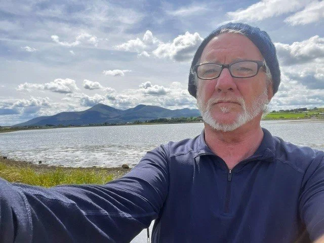 A man with glasses, a beard and a mustache taking a selfie outdoors beside a body of water with mountains in the background on a partly cloudy day.