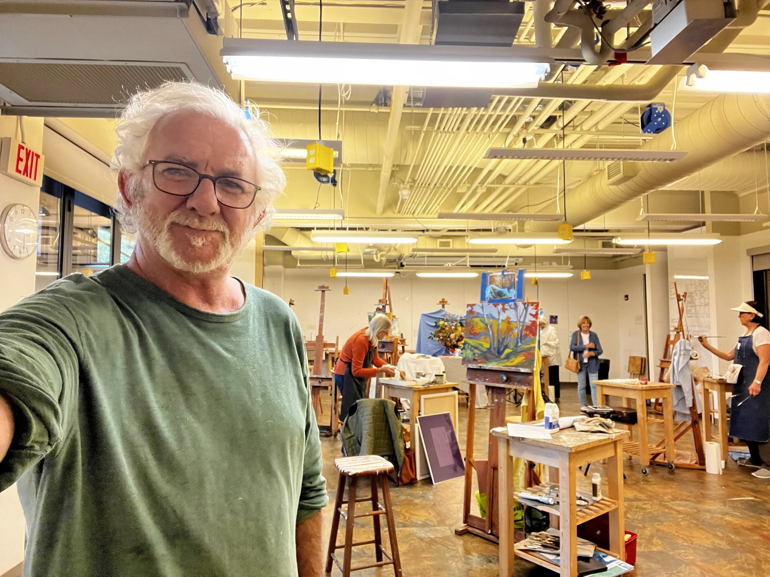 A man with glasses, white hair, and a beard taking a selfie in an art studio filled with painters and completed paintings.
