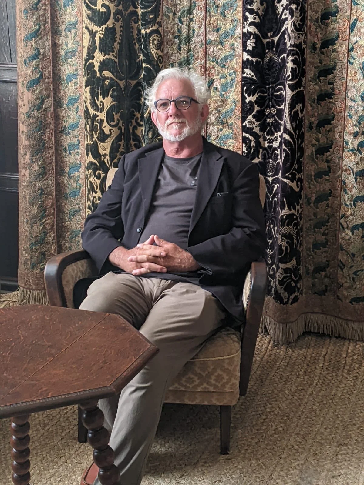 An elderly man with white hair, glasses, and a beard, seated in an ornate armchair with a backdrop of patterned curtains.