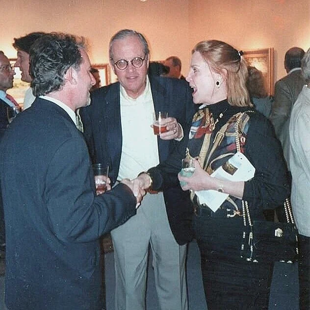 Three people at a social event conversing, two men and one woman, holding drinks, with other people in the background.