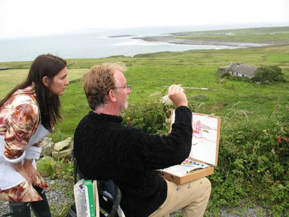 An artist painting outdoors on a grassy hill overlooking the ocean, with a woman observing.