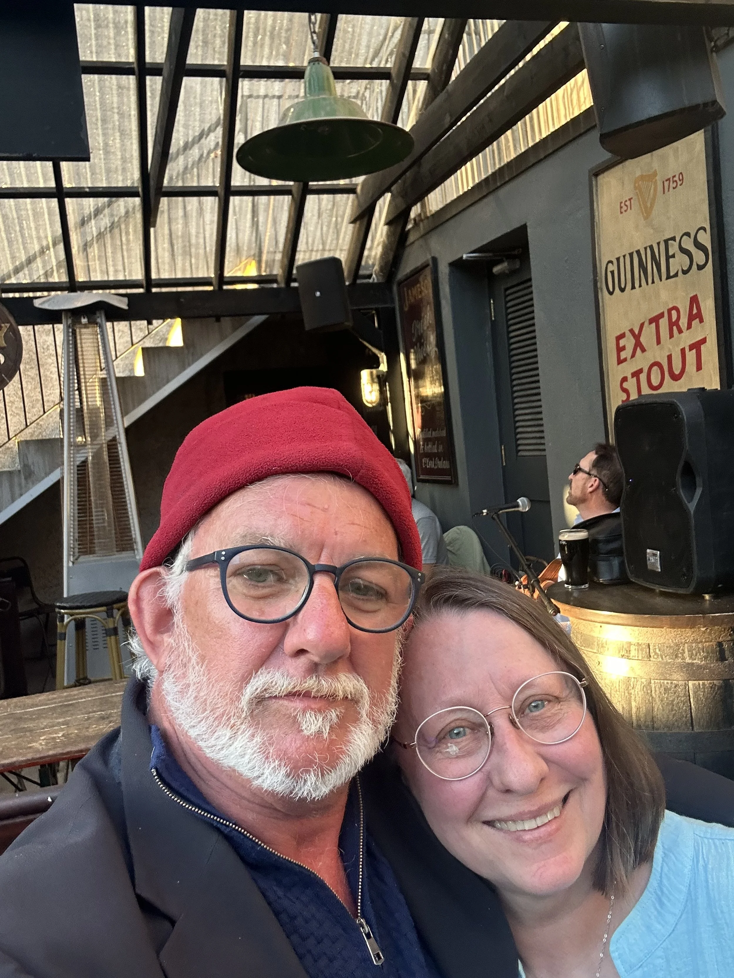 Close-up of a smiling man and woman wearing glasses, taking a selfie in a restaurant or pub with a rustic interior, including a Guinness ad and a stage with a musician in the background.