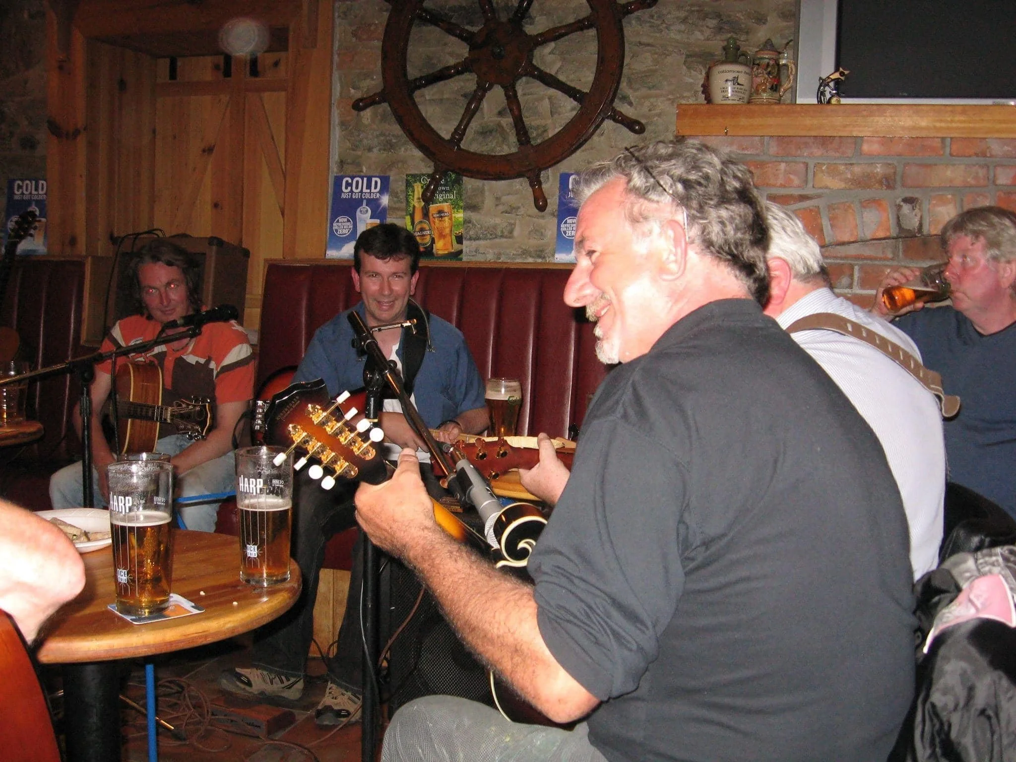 Group of men playing guitars and singing in a cozy pub with brick walls and wooden decor, some drinking beer.