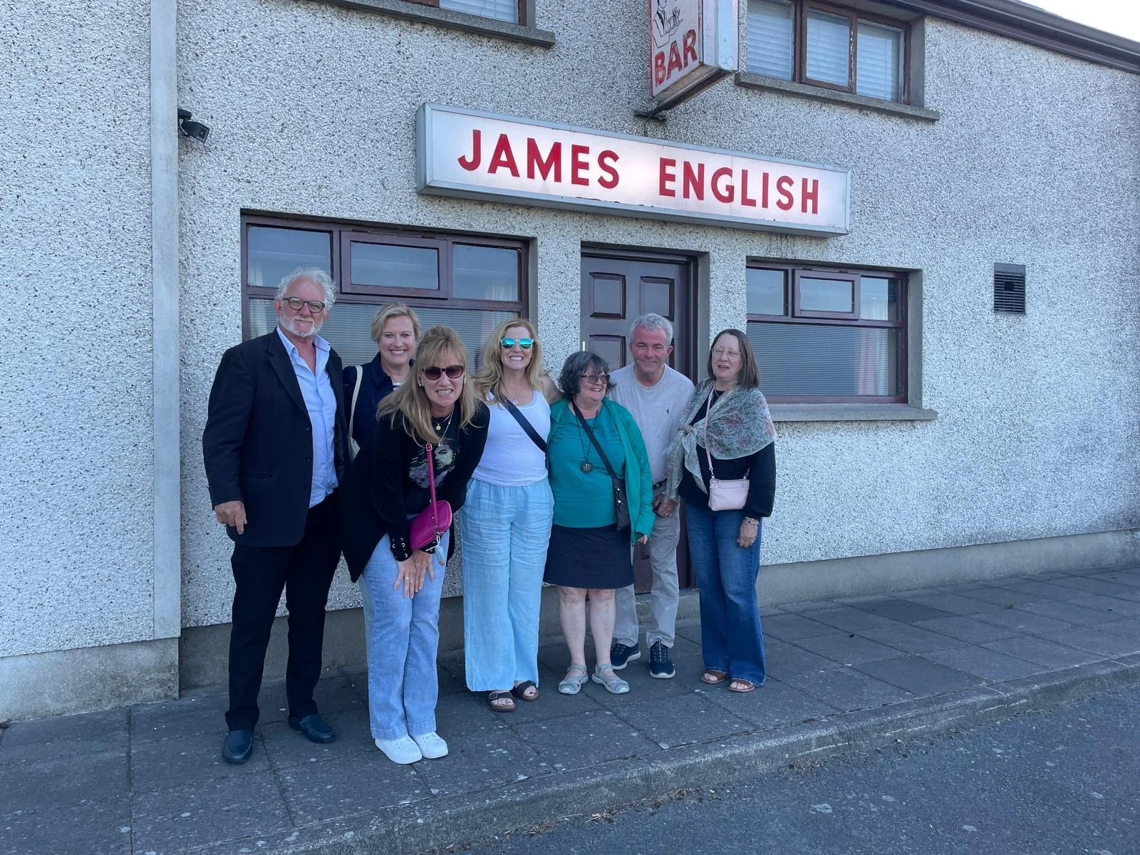 Seven people standing in front of a building with a sign that reads 'James English'. The group is smiling and posing for the photo. The building has a textured gray exterior and two windows with brown frames.