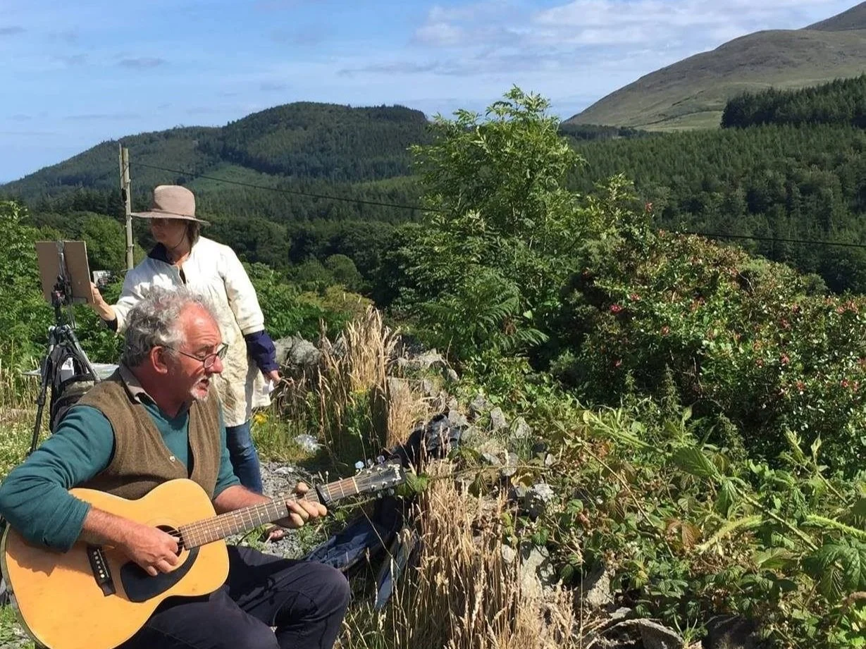An older man playing an acoustic guitar outdoors with two women nearby, surrounded by green trees and mountains under a partly cloudy sky.
