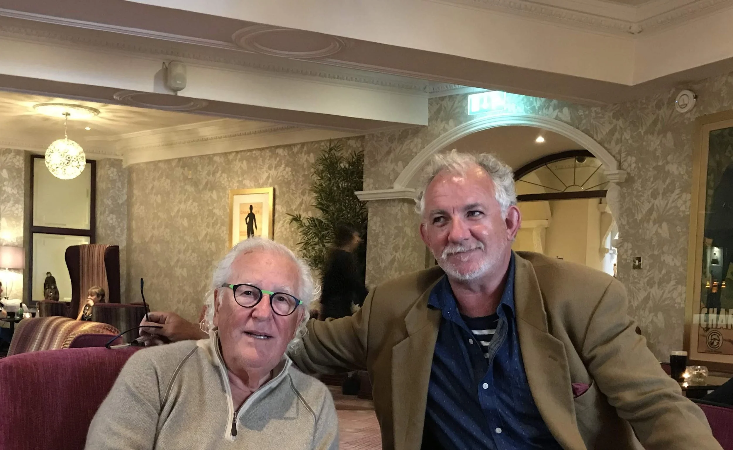 Two men sitting in a restaurant, one with glasses and white hair, the other with curly gray hair and a beard, smiling at the camera.