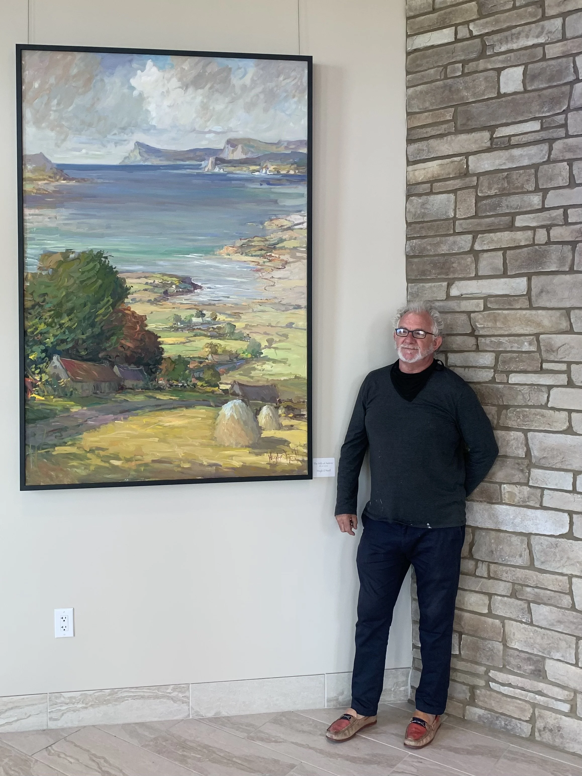 A man with white hair, beard, and glasses stands next to a colorful landscape painting in an art gallery. The painting depicts a coastal scene with water, hills, trees, and rocks.