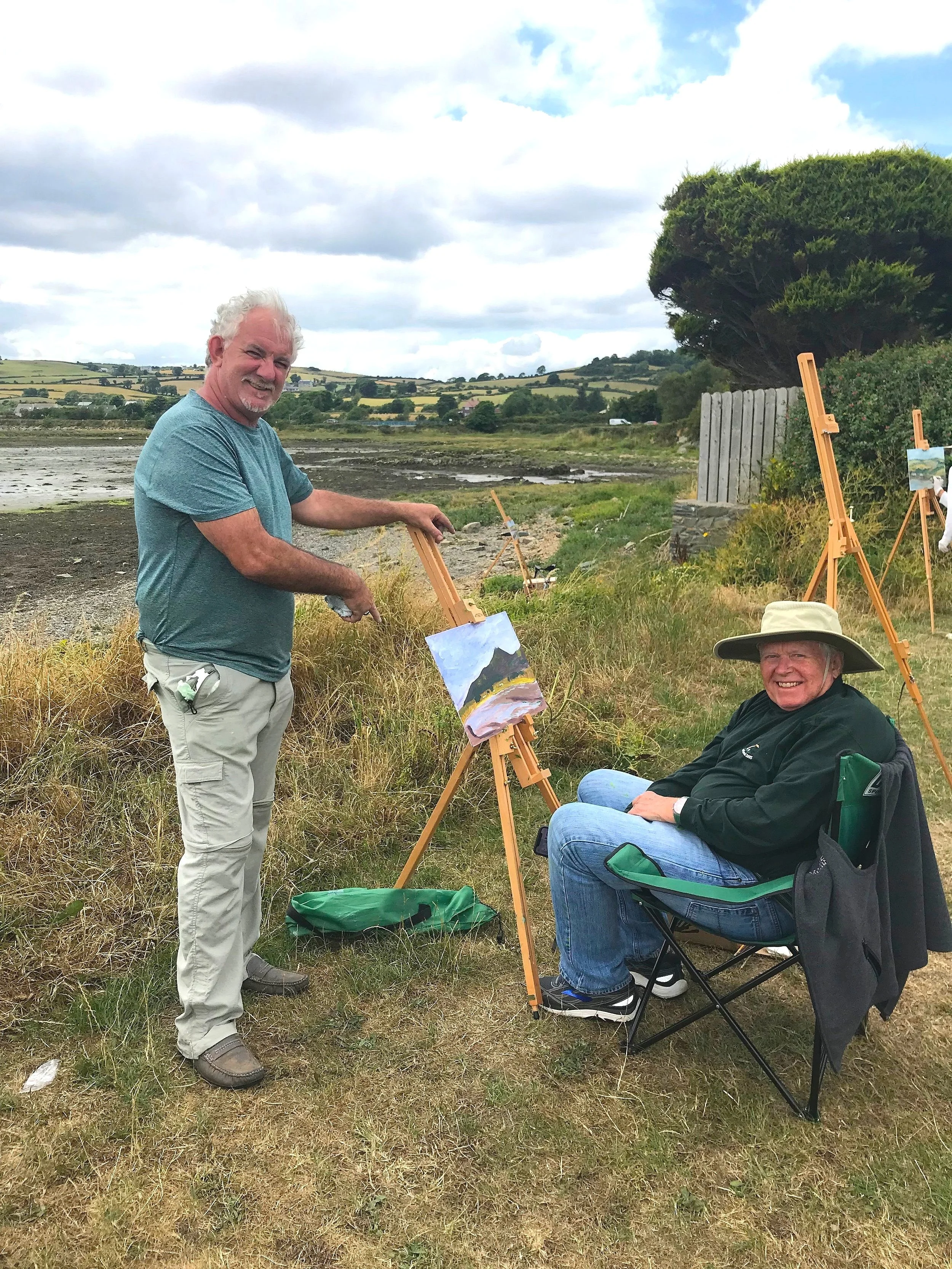Two men painting outdoors near a water body, with a landscape of rolling hills and cloudy sky in the background. One man is standing, smiling, holding a paintbrush; the other is sitting in a chair, smiling, wearing a wide hat and black jacket, with a