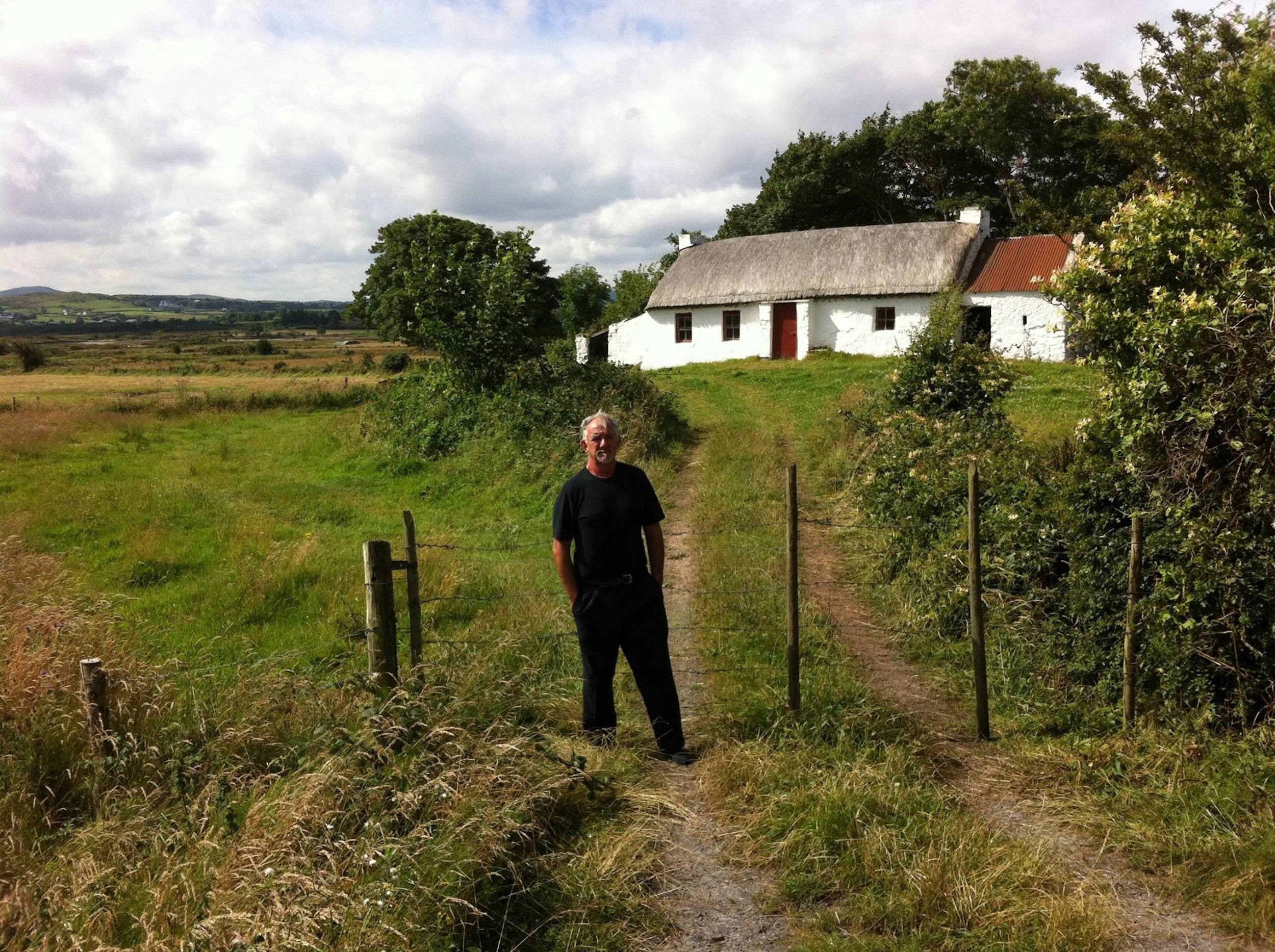 A man standing on a grassy dirt path near a white cottage with a thatched roof in a rural landscape. The sky is partly cloudy, and the scene features open fields and distant hills.