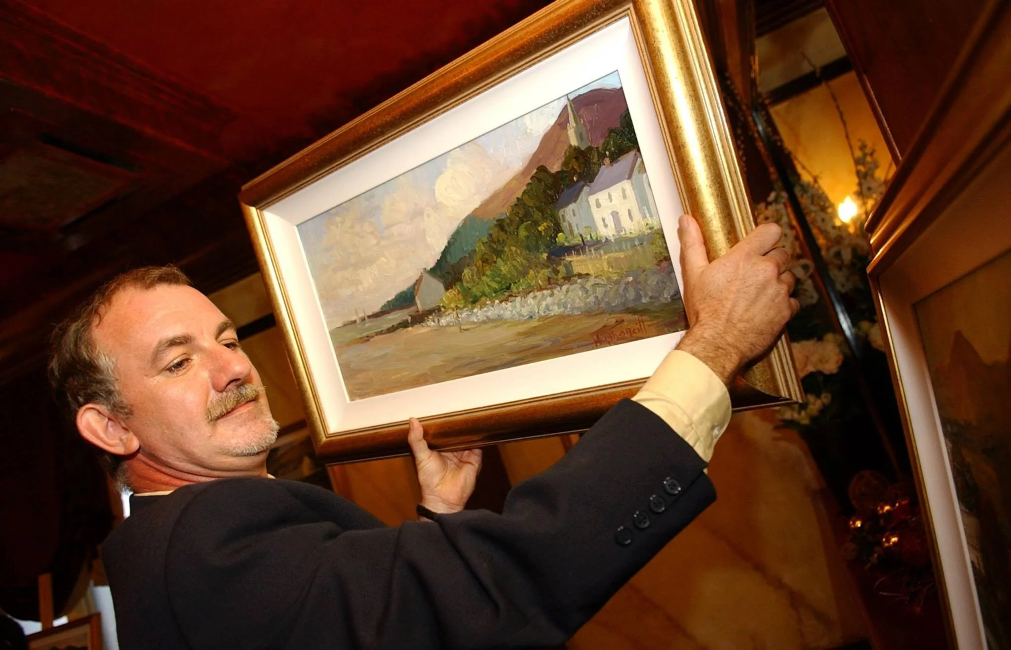 Man holding a framed landscape painting with a gold frame, inside a room with warm lighting, flowers in the background.