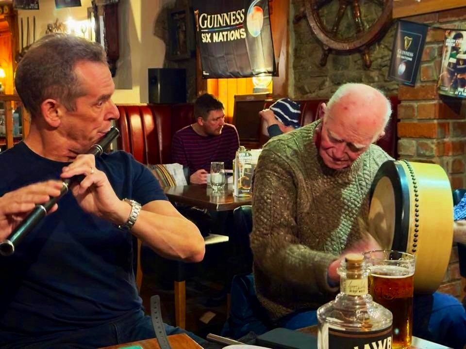 Two men playing traditional Irish music in a pub, one with a flute and the other with a bodhrán, with drinks and a Guinness banner visible.