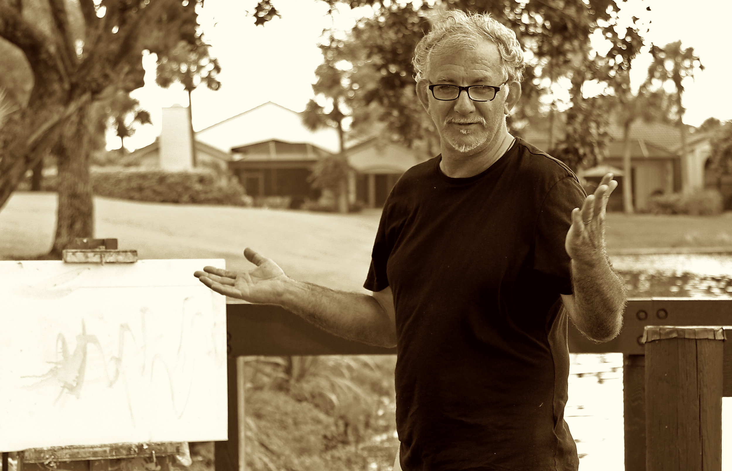 A man with glasses and curly hair, wearing a black t-shirt, standing outdoors near a body of water and a wooden bridge, gesturing with his hands and looking at the camera.