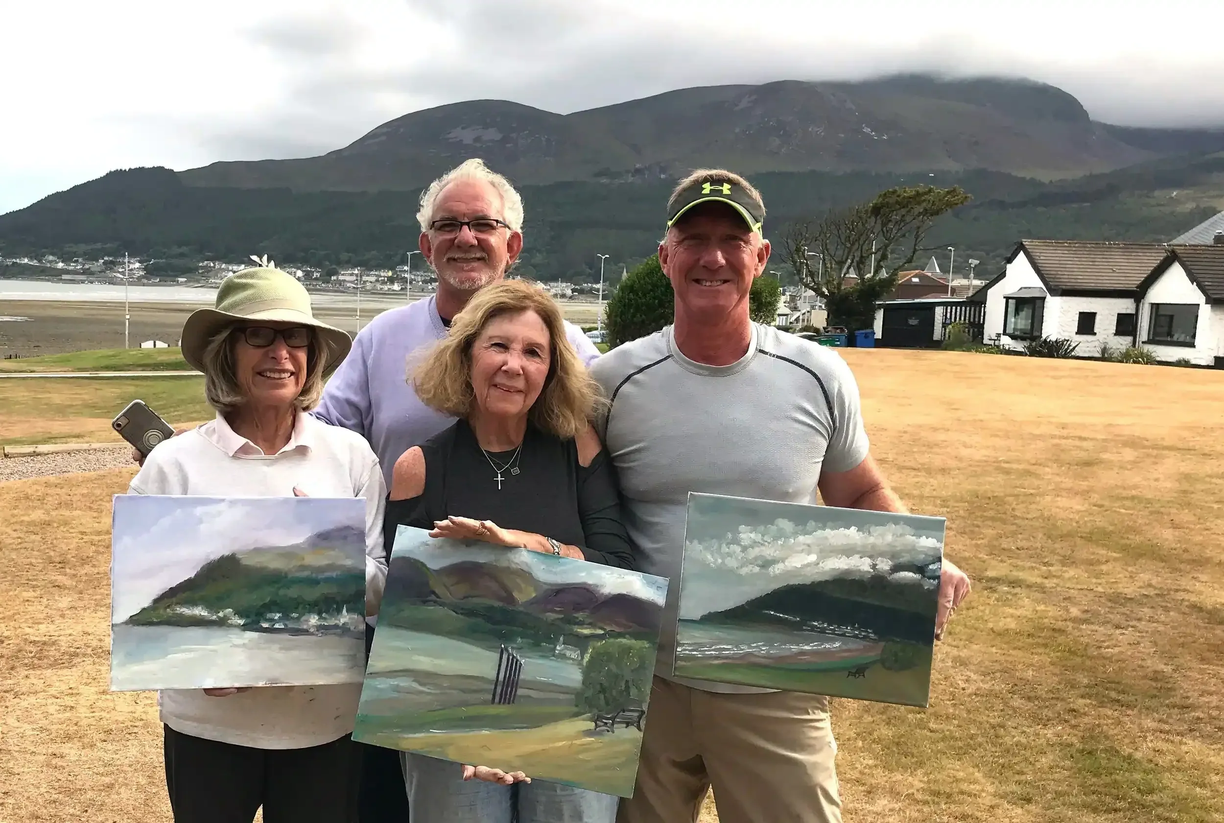 Group of five people outdoors holding landscape paintings with a mountain and houses in the background.
