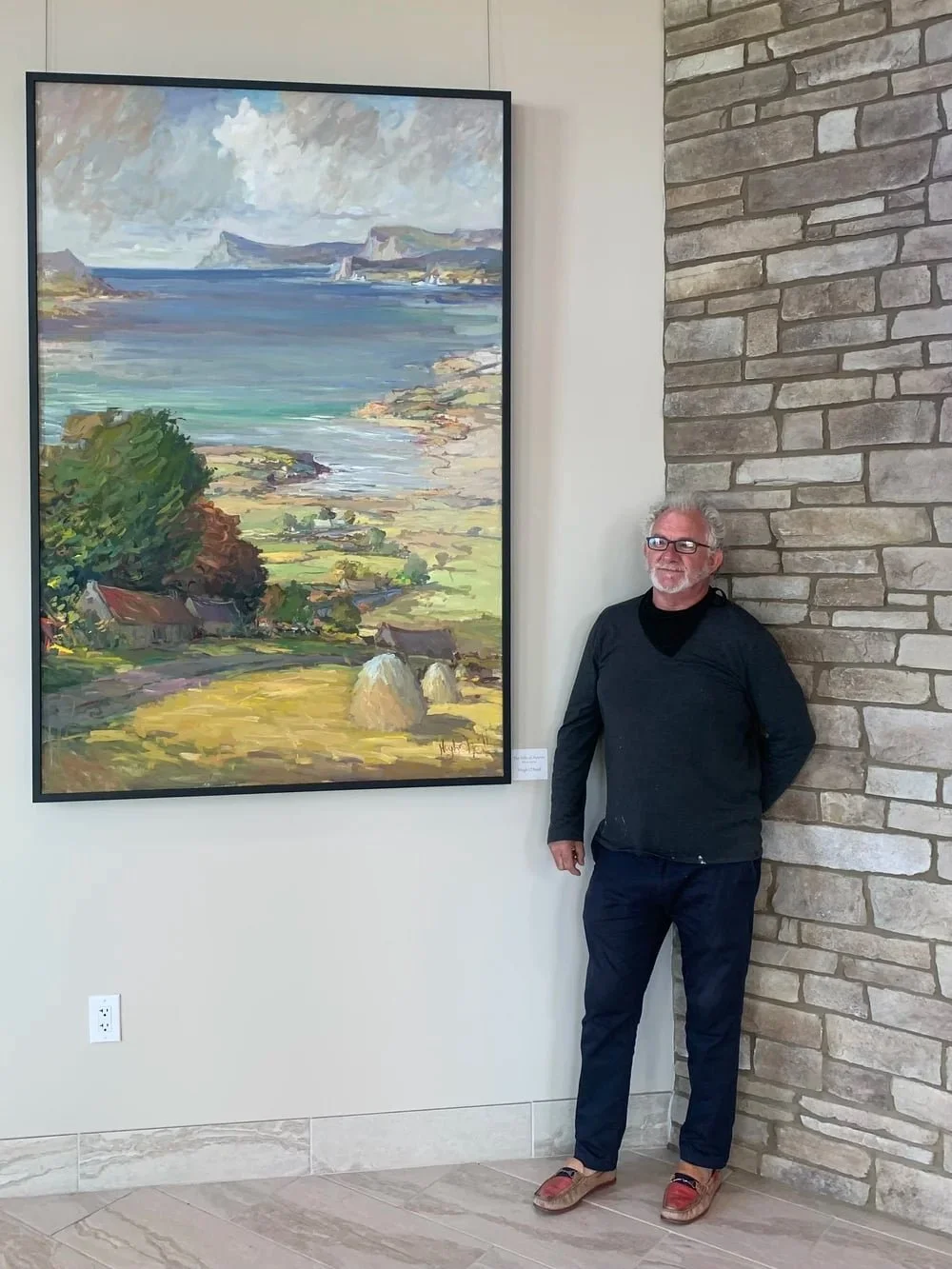 A man with gray hair, glasses, wearing a black jacket and dark pants, stands next to a large landscape painting on a gallery wall. The painting depicts a coastal scene with water, cliffs, greenery, and houses.