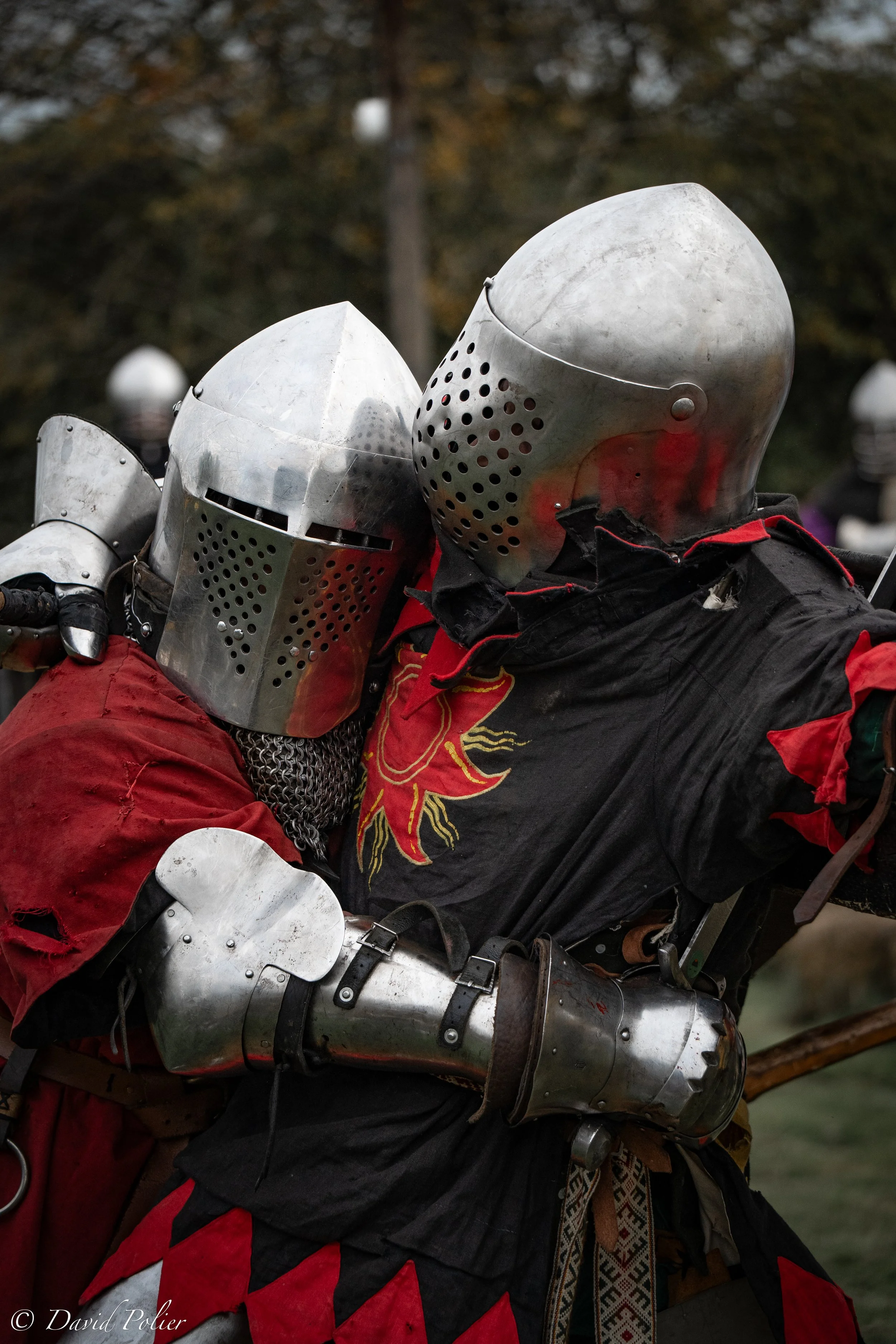 Two people dressed in medieval knight costumes embracing, wearing metal helmets and armor, at an outdoor event.